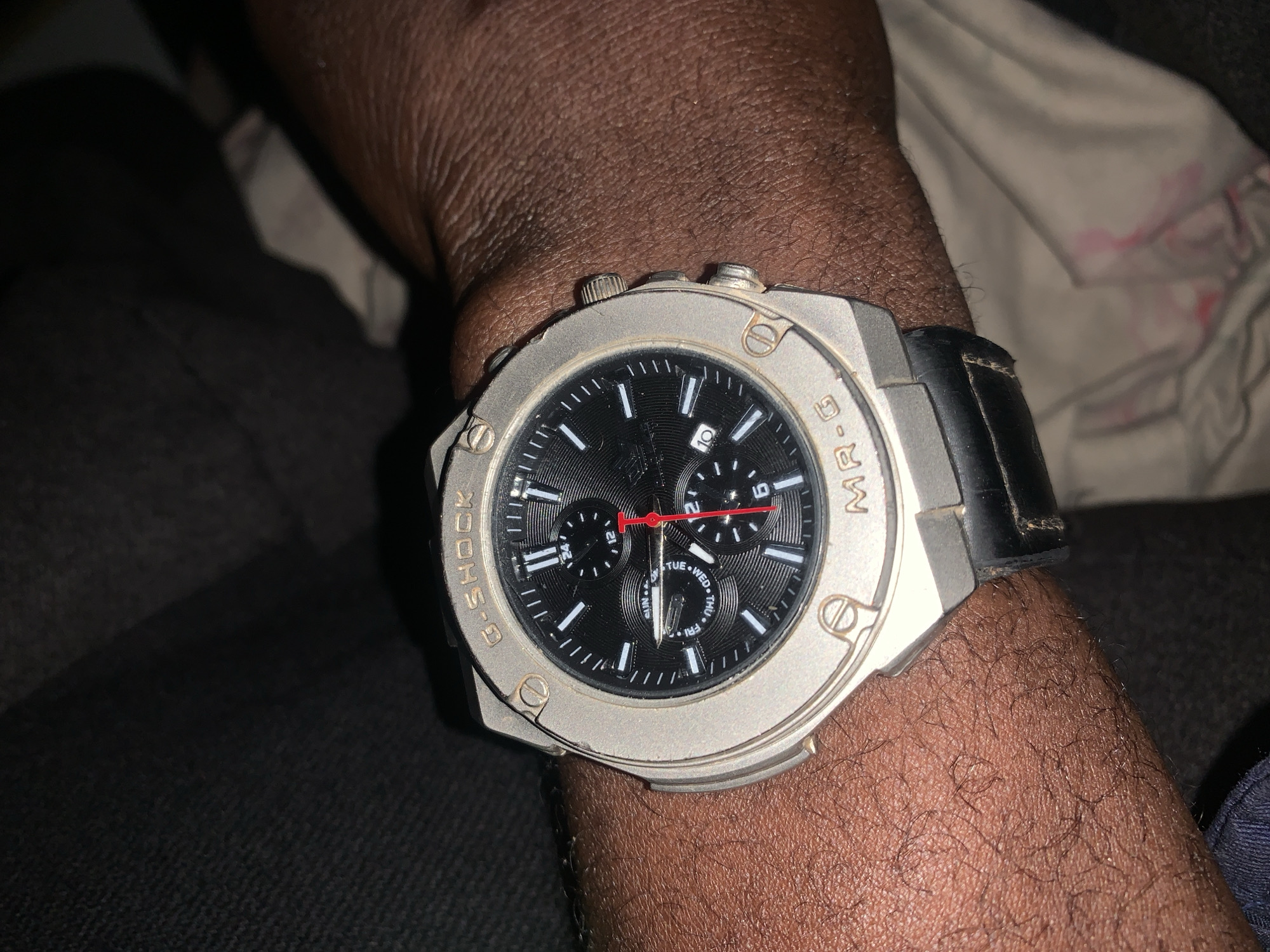 The image shows a close-up view of a person's wrist wearing a G-SHOCK MR-G watch. The watch has a silver-toned metal case and a black dial with white hour markers. There are three sub-dials and a date window. The watch band is made of black leather. The person's skin tone is dark, and there is visible hair on the wrist. The background is dark and out of focus, suggesting an indoor environment. The watch is positioned on the wrist as if being worn normally. There is no discernible indication of the city of New Stadium, Nigeria.