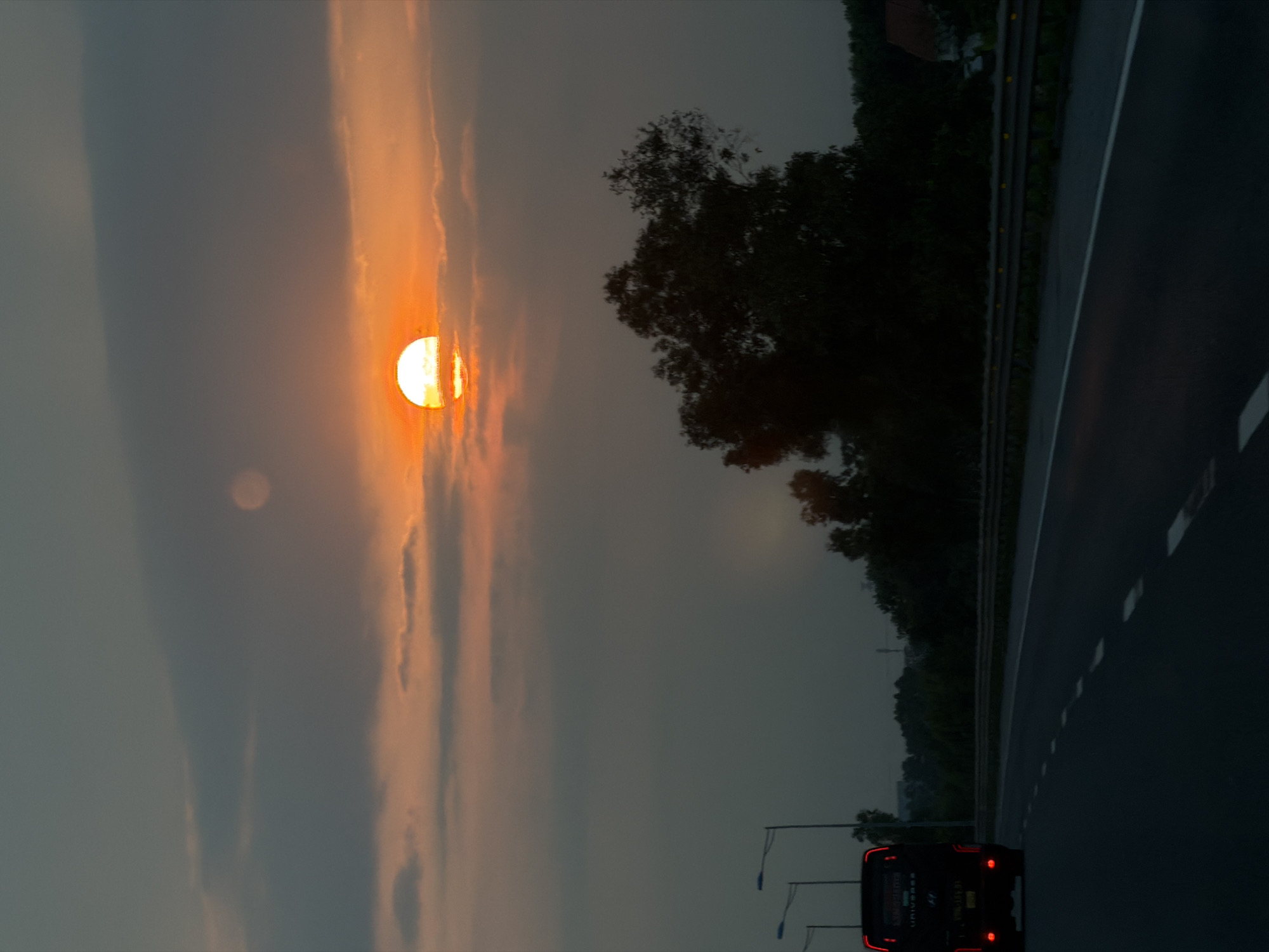 This image captures a twilight scene from a highway in Hải Phòng, Vietnam, likely at sunset. The setting sun, a large, intensely bright orange orb, dominates the left side of the frame, partially veiled by thin clouds that glow with its warmth. To the left of the sun, a fainter, circular light source is also visible in the hazy sky. The sky transitions from vibrant orange and yellow hues near the sun to a softer, muted grey-blue further away.
In the foreground, a dark asphalt highway stretches from the bottom of the frame towards the upper right, delineated by white dashed lane markings. Along the right side of the highway, a metal guardrail separates the road from a dense embankment of dark green trees, their silhouettes stark against the lighter sky.
A black vehicle, possibly a bus or large SUV, is seen in the bottom right corner, receding into the distance. Its red taillights are illuminated, standing out against the dark body. Visible text on the rear of the vehicle includes "UNIVERSE" in white lettering, suggesting its make or model. Below this, there are other smaller, indistinct symbols or words. Further down the road, two streetlights with blue hanging elements are visible along the right side, extending over the highway.
The scene conveys a sense of peaceful travel during the golden hour, with the natural beauty of the sunset illuminating the roadside landscape and the highway.