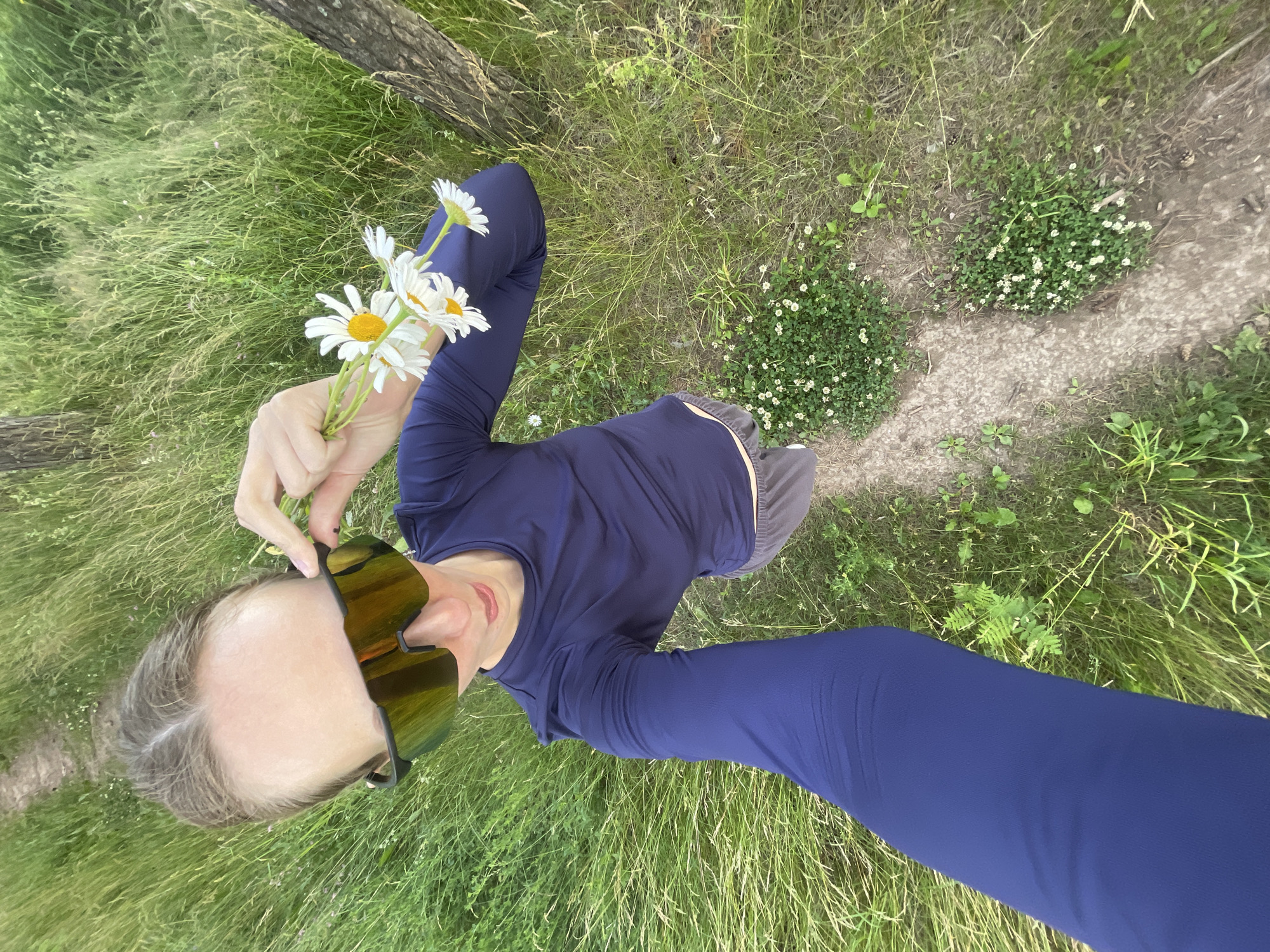The media file shows a person holding a small bouquet of daisies in their left hand. The person is wearing a long-sleeved navy blue shirt and grey shorts. They are also wearing large, mirrored sunglasses with yellow lenses. The image is taken from a high angle, looking down at the person.
The scene is an outdoor setting with tall green grass and a dirt path. There are some small white flowers growing in patches near the path. A tree trunk is partially visible on the left side of the image. The lighting suggests it is daytime, possibly late afternoon due to the warmth of the sunlight reflected in the sunglasses. The overall atmosphere appears relaxed and natural. There is no visible text in the image. The location context provided is Podshivalovo, Russia.