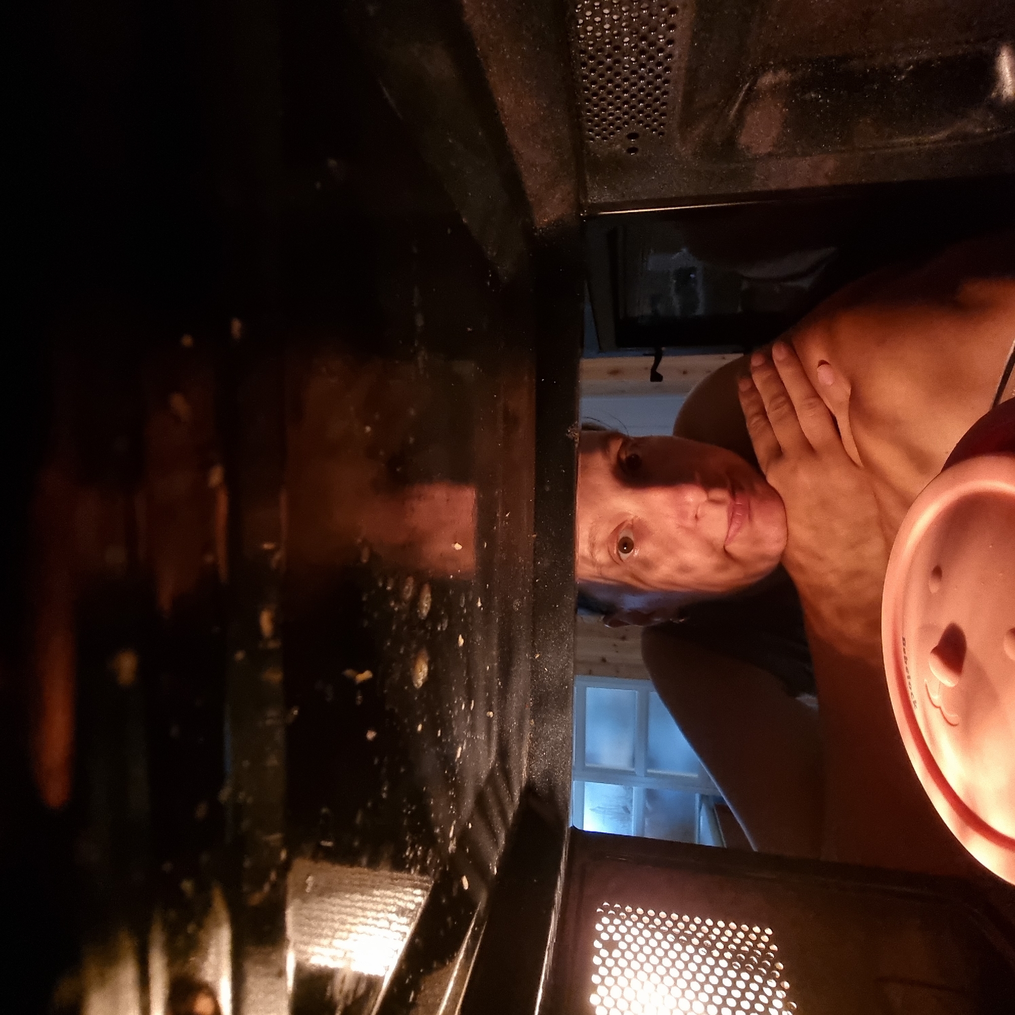 An overhead, interior shot of a microwave shows a person inside with their head resting on their arms. The person's face is visible, looking directly at the camera. In the foreground, a pink food container with a lid is partially visible. The interior of the microwave appears dirty with splatters on the ceiling. The background shows a window with a frosted glass pane and wooden paneling, suggesting the microwave is located in a kitchen. The scene is lit by the microwave's interior light and the ambient light from the window. No specific location in Dunnae-myeon, South Korea can be determined from the image.