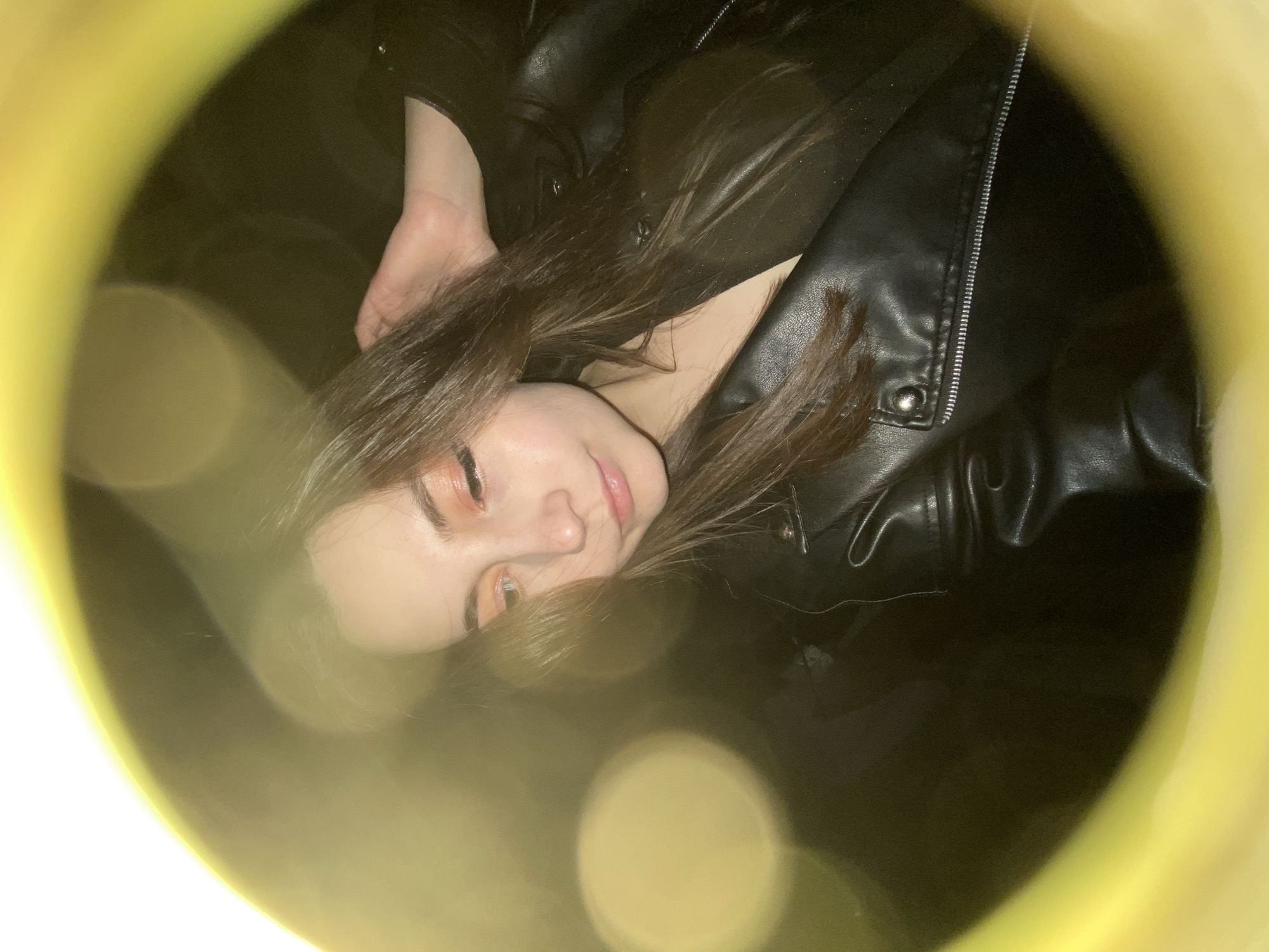 A woman lies on her back, wearing a black leather jacket. Her brown hair is partially obscuring her face. She has fair skin and wears makeup, including eye shadow and lip color. The scene is framed by a circular, blurred yellow border, creating a sense of looking through an object. There is no information to suggest it was taken in Chaikovsky, Russia.