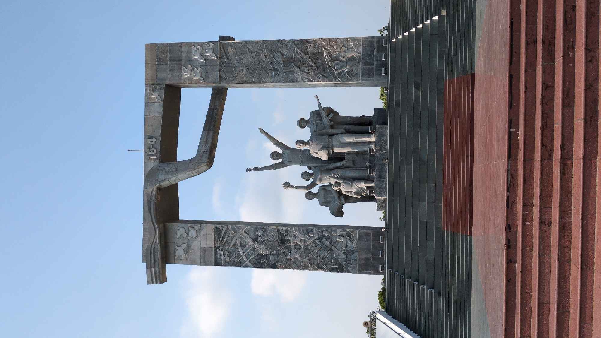 A horizontal shot depicts a monument located in Phan Rang – Tháp Chàm, Vietnam. The monument features a sculptural group of figures. These figures are placed within a gray stone structure that includes a depiction of "16-4". The stone structure has a relief carving showing individuals wielding weapons. Steps of red and gray stone are present to the right of the sculptural group. The background features a clear blue sky.