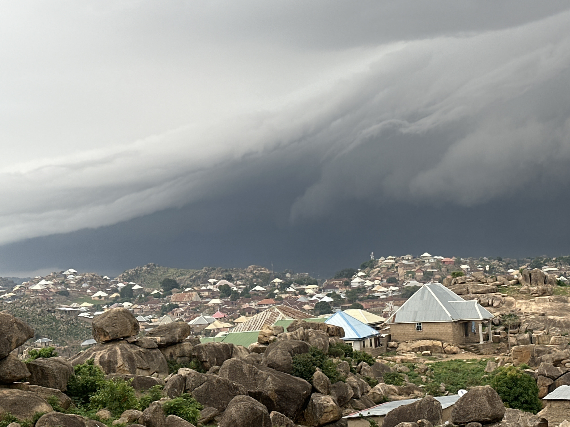 The image depicts a densely populated area within Jos, Nigeria, characterized by closely packed, mostly single-story dwellings scattered across a hilly terrain dotted with large rock formations. The houses exhibit a variety of roofing materials and colors, including corrugated iron sheets in shades of grey, green, blue, and red. These structures are nestled amongst the natural rocks, showcasing an integration of human habitation with the existing landscape. In the foreground, a cluster of boulders dominates the view, interspersed with patches of greenery. Overhead, a dark and ominous cloud formation looms large, suggesting an impending storm. The sky is overcast, casting a somber light across the scene.