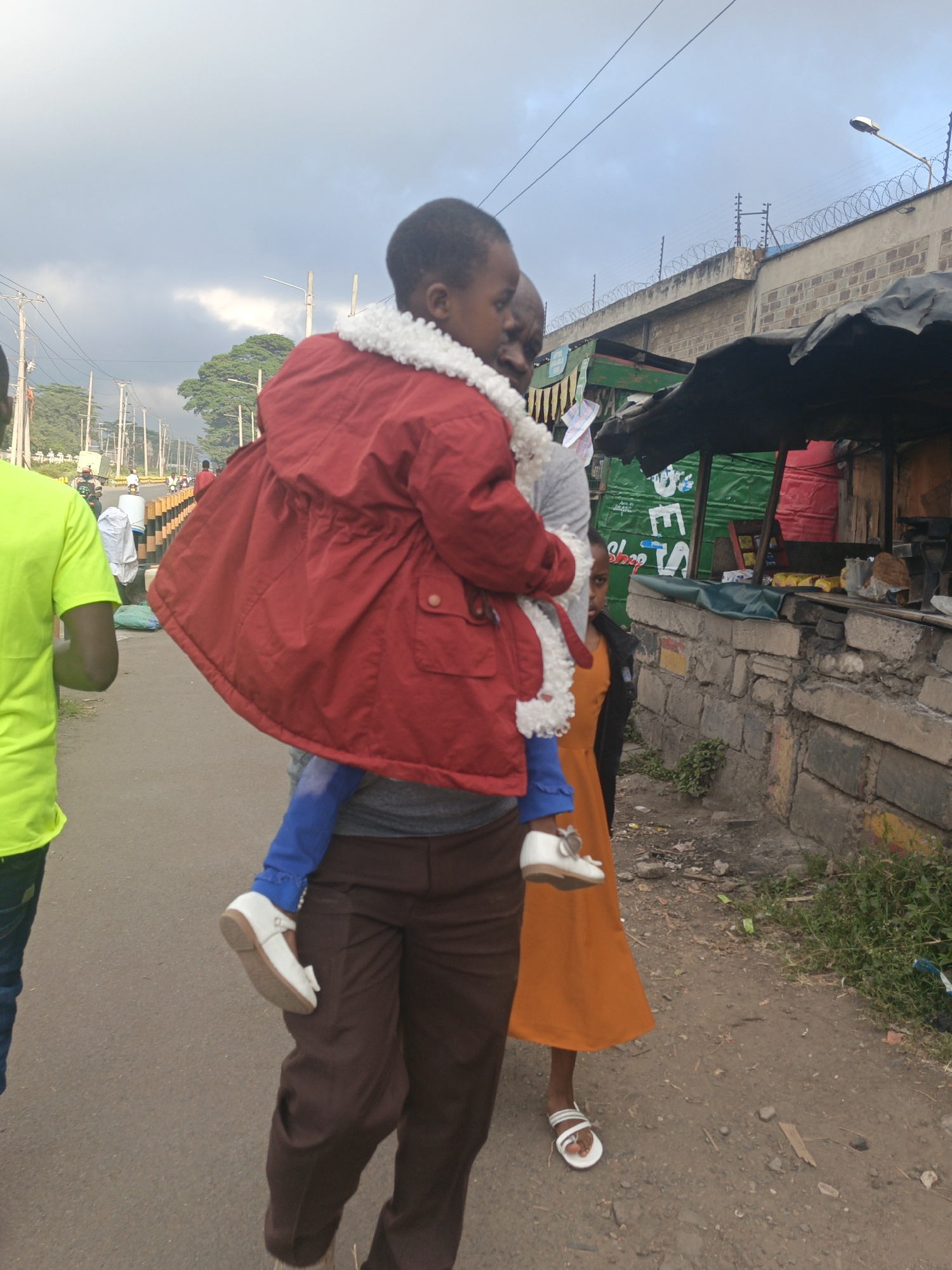 A man carries a child wearing a red coat with a white fleece collar on his back, walking on a road. He is wearing brown trousers. Another child in an orange dress walks beside him. A person in a bright yellow shirt is walking to the left of the frame. In the background is a street scene with buildings, power lines, and various shops or stalls, one of which appears to have the word "Shop" written on it. The upper background features a wall topped with barbed wire. The scene is outdoors, and the sky is overcast. The location is Nairobi, Kenya.