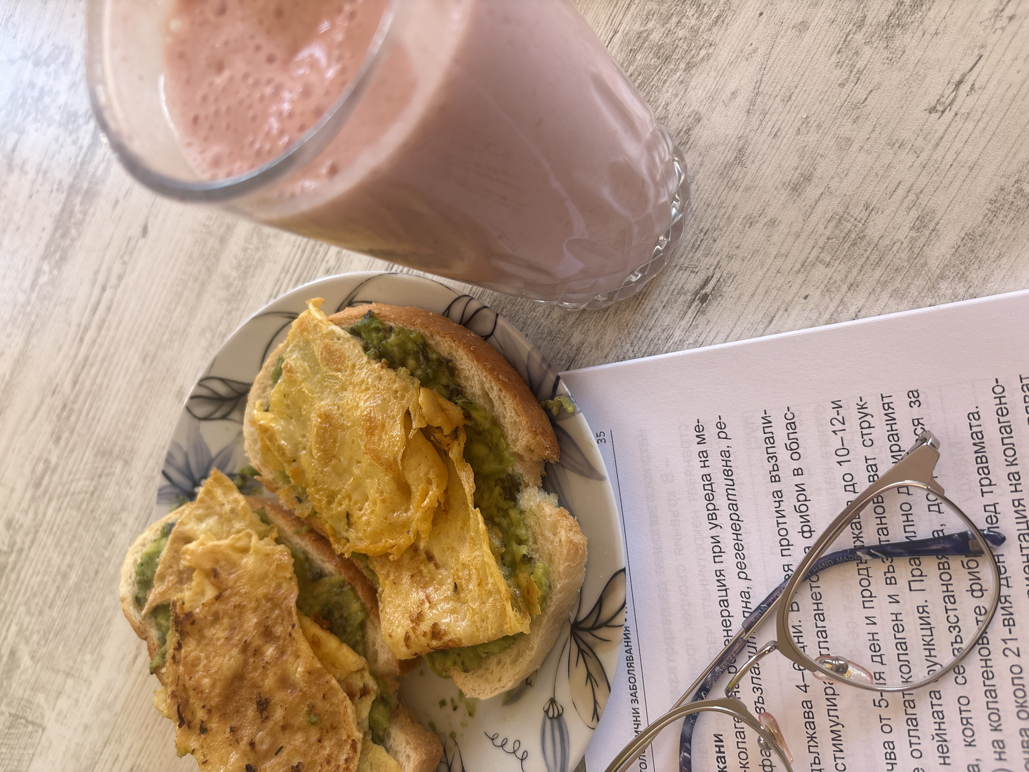 A breakfast scene. A pink smoothie in a glass, avocado toast with what appears to be scrambled eggs, and a piece of paper with text on it rest on a light woodgrain surface. On the paper lies a pair of eyeglasses. The text appears to be in Bulgarian. The image does not provide specific information regarding the location in Varna, Bulgaria.