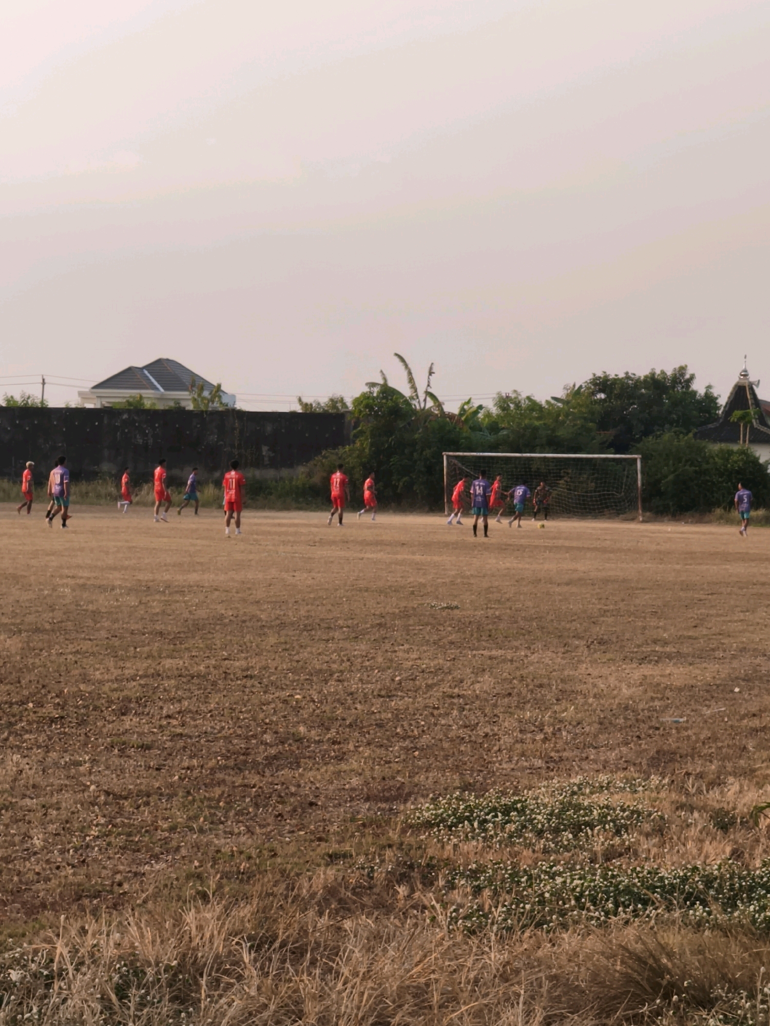 A soccer game is underway on a dry, brown field in Desa Sindon, Indonesia. The players are wearing red and blue uniforms. The field is surrounded by trees and a dark wall. In the background, there are buildings and a clear sky. A soccer goal is visible on the right side of the field. The ground is covered in dry grass and some small green plants.
