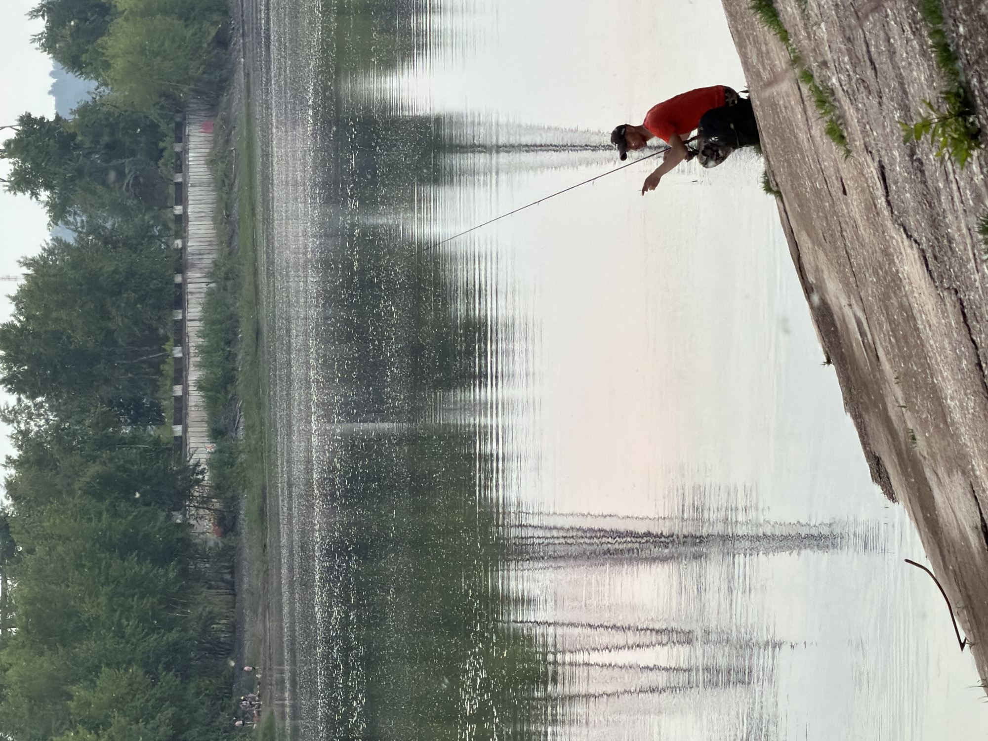 The image shows a man fishing on the bank of a body of water, likely the Kama River in Chaikovsky, Russia. The man is wearing a red t-shirt and a dark baseball cap, and he is crouched down with a fishing rod. His fishing rod is extended over the water. He has a backpack with a camouflage pattern next to him. The bank is a steep, sloped surface of concrete or rock, with some sparse vegetation growing on it. In the background, across the water, there is a dense line of trees and some structures that appear to be part of a city or industrial area. The water's surface is calm with reflections of trees and structures. There are small groups of people visible on a distant bank, and more trees and buildings are visible in the far background. The sky is overcast.