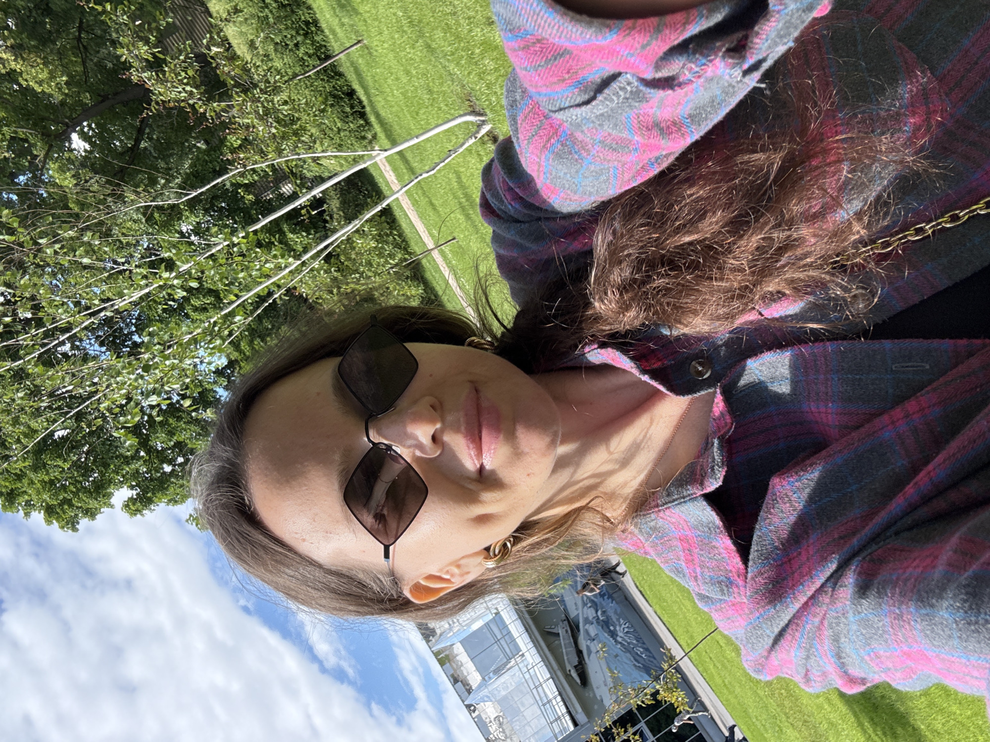 A woman with brown hair and sunglasses takes a selfie outdoors. She is wearing a pink and grey plaid shirt. In the background, there is a large modern building with a reflective facade, grassy areas, and trees. The sky is blue with white clouds. The location is the Moscow State University campus in Moscow, Russia.