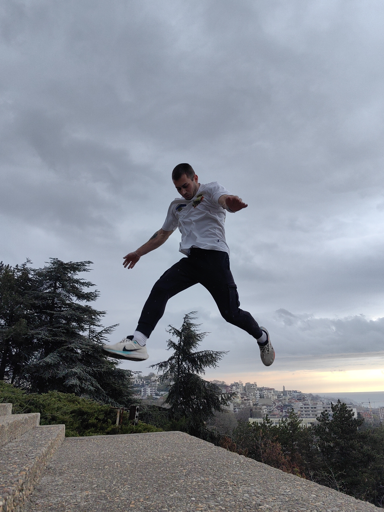 A full-length shot captures a man mid-jump against a cloudy sky. The man is wearing a white t-shirt, black pants, and sneakers. He is positioned in mid-air, seemingly leaping from a concrete surface at the edge of a hill.
In the background, there is a view of Varna, Bulgaria, including buildings, trees, and the Black Sea. Trees surround the concrete path the man jumped from. There are stairs to the left of the path, which appears to be made of concrete. The overall atmosphere suggests an overcast day near dusk.