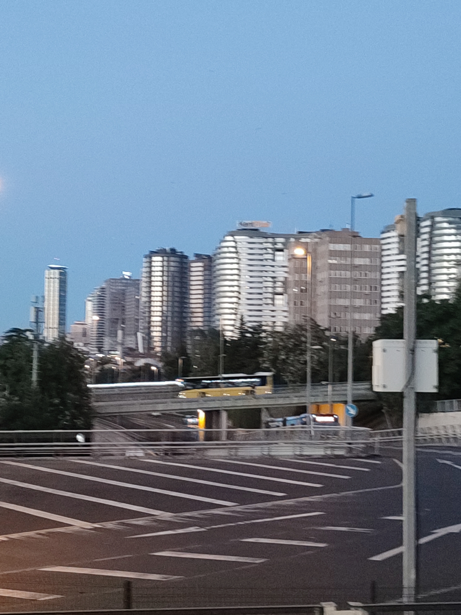 In Kadıköy, Turkey, a cityscape view captures a cluster of high-rise buildings against a twilight sky. A prominent yellow bus is visible on an elevated roadway. The foreground shows a street with white lane markings. The overall scene suggests an urban environment at dusk.