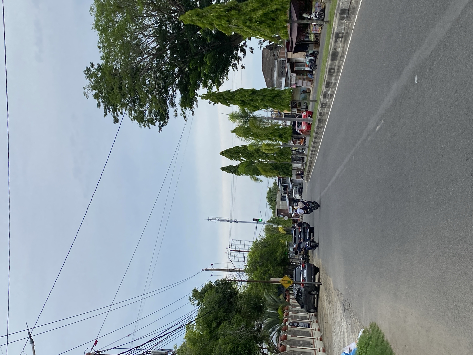 The image depicts a street scene in Lhokseumawe, Indonesia, likely on Jalan Malikussaleh. The viewpoint is from a slightly elevated position on the side of the road, looking down the street. The sky is overcast with a pale blue and white hue. On the left side of the road, a white fence with decorative pillars lines the property. Several vehicles are visible: a dark gray SUV is parked on the left shoulder, and several motorcycles and a few cars are moving along the road in both directions. A traffic light with a green signal is visible in the distance, suspended above the intersection. Numerous tall, slender green trees are planted along the roadside, alternating with palm trees. Buildings with tiled roofs are visible behind the trees and shops. The road itself is asphalt with white lane markings. Power lines crisscross the upper portion of the frame, supported by utility poles. The overall impression is of a moderately busy urban street in Lhokseumawe on a cloudy day.