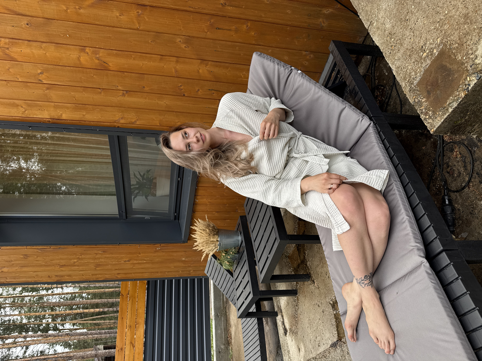 The image shows a woman in a white bathrobe lying on a gray chaise lounge, on a patio or deck. The woman is facing the camera and her legs are crossed. Ankle tattoo visible on the left leg. There is a small black side table with a bucket containing wheat or reeds next to the chaise lounge. The backdrop includes a wooden wall with a window. The setting appears to be outdoors, possibly a resort or private property. The precise location within the city of Chaykovsky, Russia, is not discernible from the image.