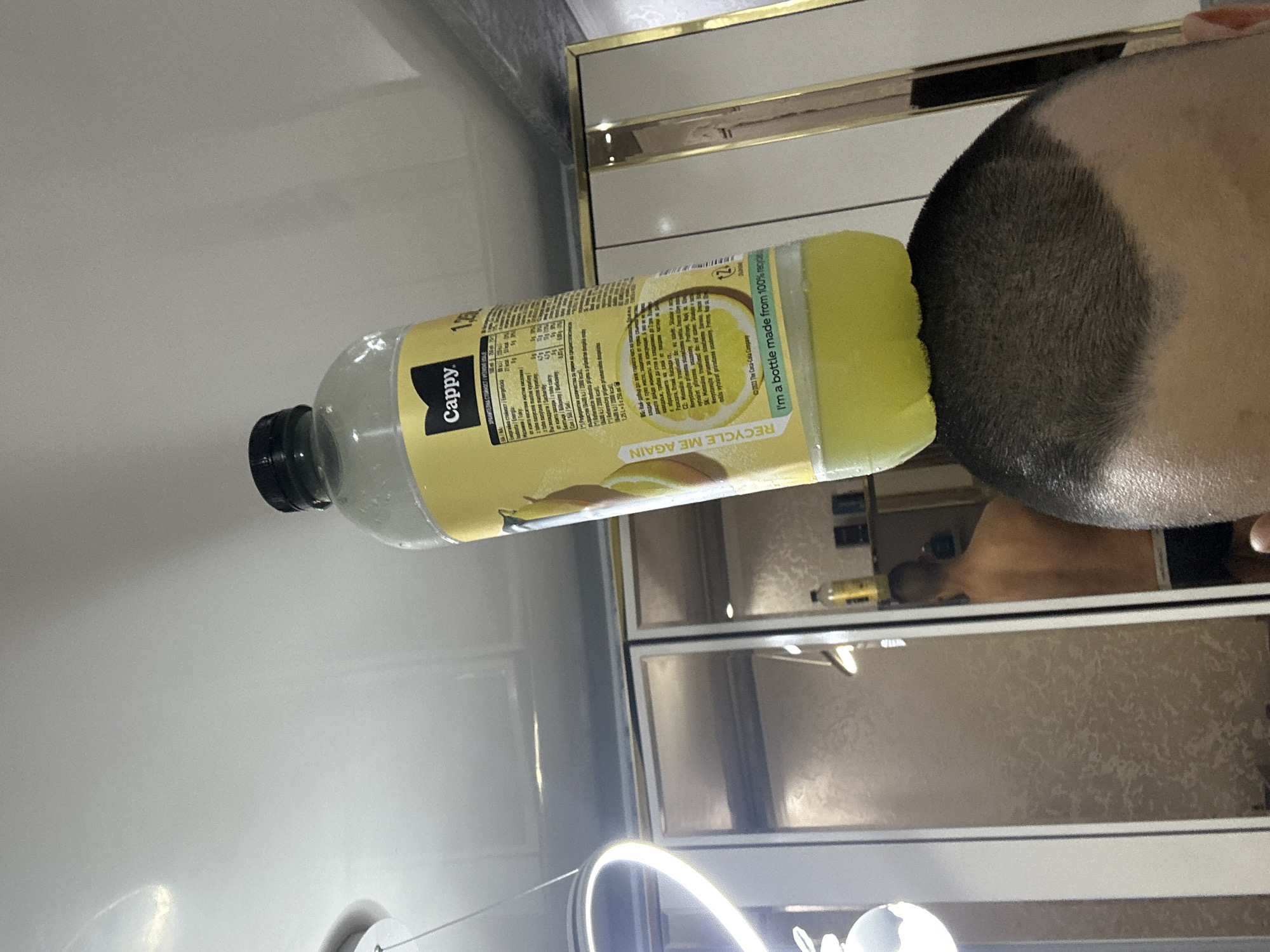 An indoor, vertically oriented close-up shot features a plastic bottle of Cappy brand lemonade held upright, with the base of the bottle partially obscuring the top of a person's head. The bottle contains a light yellow liquid, and its black cap is visible at the top left. The main label on the bottle is yellow and contains nutritional information and brand text.
The person, whose head is mostly visible on the right side of the frame, has short, dark hair with a faded cut. The reflection of this person, seen from behind with bare shoulders, is visible in mirrors or reflective panels in the background. These reflective surfaces also show a room interior, possibly a bathroom or dressing area, with golden or metallic frames. A bright, circular light fixture is prominently visible in the lower foreground, suggesting artificial illumination.
Visible text on the bottle includes "Cappy.", "1.25L", "RECYCLE ME AGAIN", "I'm a bottle made from 100% recycled plastic", and "© 2023 The Coca-Cola Company." Nutritional information in various languages, including Bulgarian ("ХРАНИТЕЛНА СТОЙНОСТ" - Nutritional Value), is also discernible on the label. The overall setting appears to be a well-lit interior, possibly at night given the reliance on artificial light. No specific activity beyond posing for the photograph is evident.