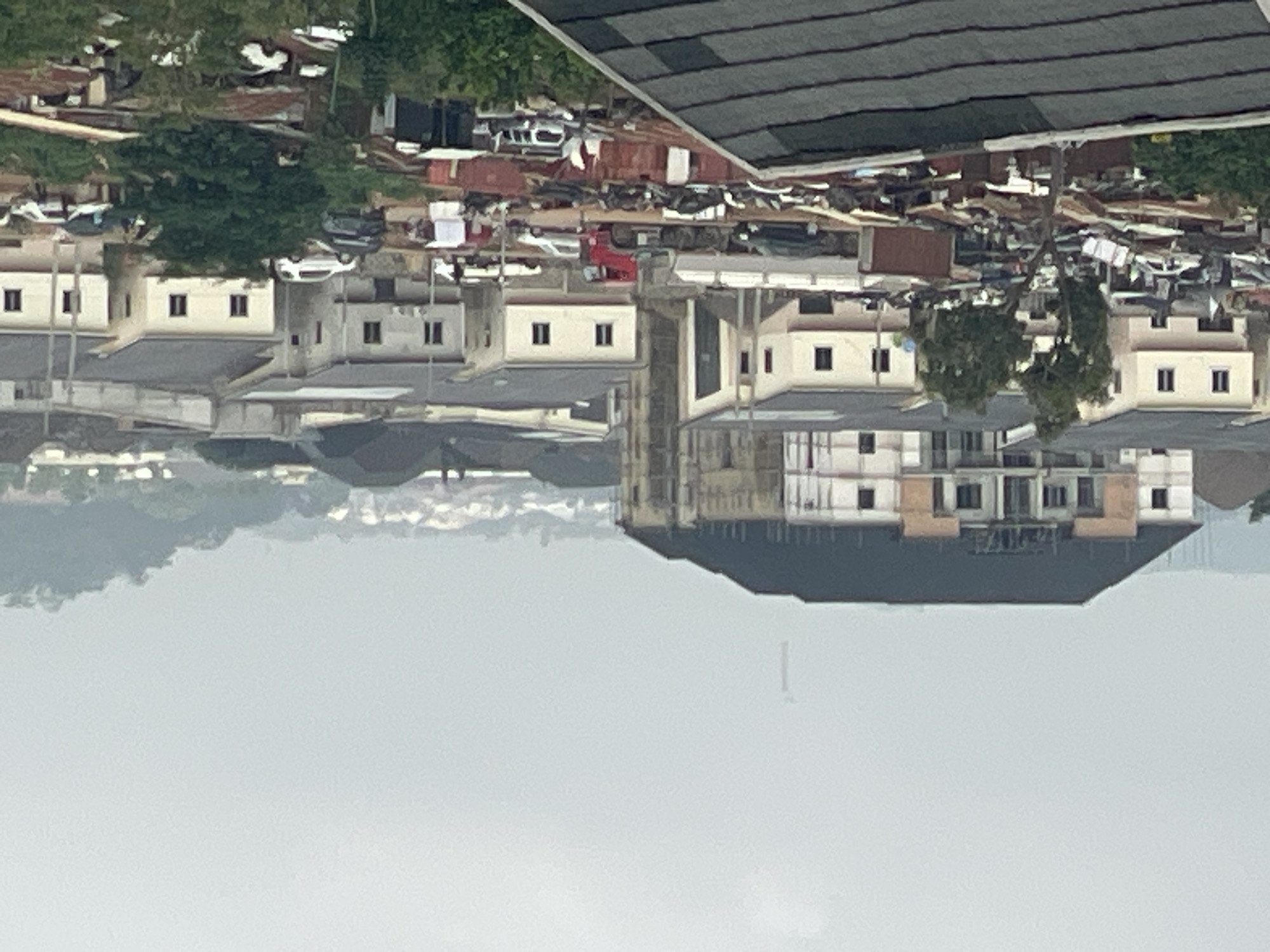 The image depicts a dense urban landscape in Abuja, Nigeria, characterized by a mix of residential and commercial structures, undeveloped land, and informal settlements. In the foreground, a dark, angled roof of a building is visible. Beyond this, a sprawling area of informal structures with corrugated iron roofs and miscellaneous debris creates a cluttered, low-income settlement. Several vehicles, including cars and what appears to be a truck, are scattered throughout this area.
Moving further into the scene, a row of modern, multi-story residential buildings with white facades and dark roofs stands out. These buildings appear to be under construction or newly built, with some showing exposed structural elements. Trees and vegetation are interspersed among these buildings.
In the mid-ground, a large, unfinished multi-story building dominates the left side of the frame. Its facade is a mix of concrete and plaster, with dark window frames and a dark, sloped roof. A red truck is positioned on a road in front of these newer buildings, suggesting ongoing activity or transport. Several other vehicles are visible on the roads and in parking areas.
The background reveals more of the city's sprawl, with numerous other buildings and residential areas extending towards rolling hills covered in vegetation. The sky is overcast, casting a muted light over the entire scene. A faint, tall, industrial-looking structure is visible in the far distance against the hazy sky, potentially a radio mast or a similar tower. The overall impression is one of rapid urban development coexisting with older, less formal areas.