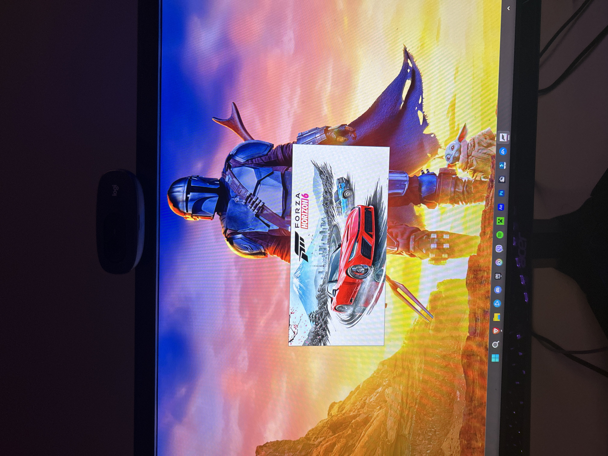 The image shows a computer monitor displaying a background featuring the Mandalorian and Grogu. An open window displays the box art for Forza Horizon 6. A black Logitech mouse is visible to the left of the monitor. The monitor's taskbar is visible, showing several application icons. The scene is indoors with dark surroundings. There is no indication that the image was taken in Burgas, Bulgaria.