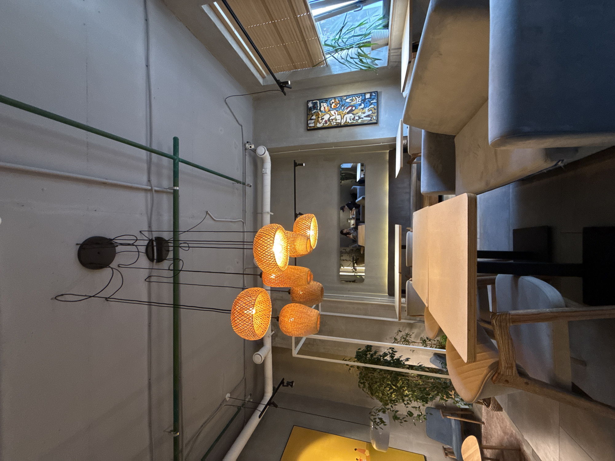 This is a wide-angle, interior shot of a modern, well-lit space, possibly a cafe or restaurant, in Khimki, Russia. The scene is depicted from a high vantage point looking downwards, with a significant portion of the image rotated. The dominant features are the clean, concrete walls and ceiling, creating a minimalist aesthetic.
Suspended from the ceiling are several woven pendant lights with a warm, orange glow, suggesting it is likely evening or the interior lighting is intentionally dim. A green pipe runs horizontally across the left side of the frame, adding a touch of industrial design. On the wall, near the lights, are two circular wall mounts.
Further into the space, a colorful abstract painting hangs on the wall, depicting human-like figures. Above the painting, a skylight with a bamboo blind allows natural light to filter in, hinting at the time of day. To the right, plush, light-colored seating is arranged in a modular fashion, forming a curved sofa or seating area. A section of a wooden table is visible in the foreground.
In the background, a reflection in a mirror reveals more of the interior, including glimpses of what appear to be people and additional seating. Green foliage from potted plants is visible, adding a natural element to the urban setting. The overall impression is one of a contemporary and inviting environment. No visible text is present in the image.