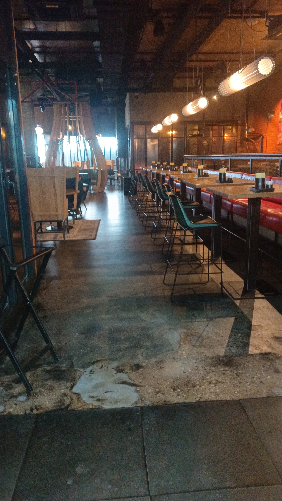 This image depicts the interior of a restaurant or bar. The scene is dominated by rows of tables and chairs, arranged for dining and drinking. The lighting appears to be dim and atmospheric, with modern tubular light fixtures hanging from the ceiling. The flooring is a dark, textured material, possibly concrete or a similar industrial finish, with some areas appearing worn or stained. There are also booths with red seating visible along one side of the room. The overall aesthetic suggests a contemporary, possibly casual dining establishment. There are no people visible in the image. The location is Mytishchi, Russia.