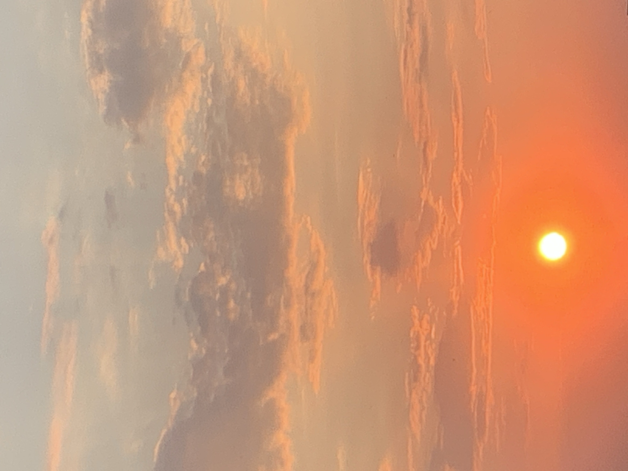 The image shows a sunset over the city of Kolomyia, Ukraine. The sky is filled with clouds that are tinged with orange and pink hues. The sun, a bright orange ball, is low on the horizon, casting a warm glow over the scene. The lower part of the image is obscured by a dark, blurry area that suggests a foreground, possibly buildings or the ground in Kolomyia. No people or specific objects are discernible in the scene.