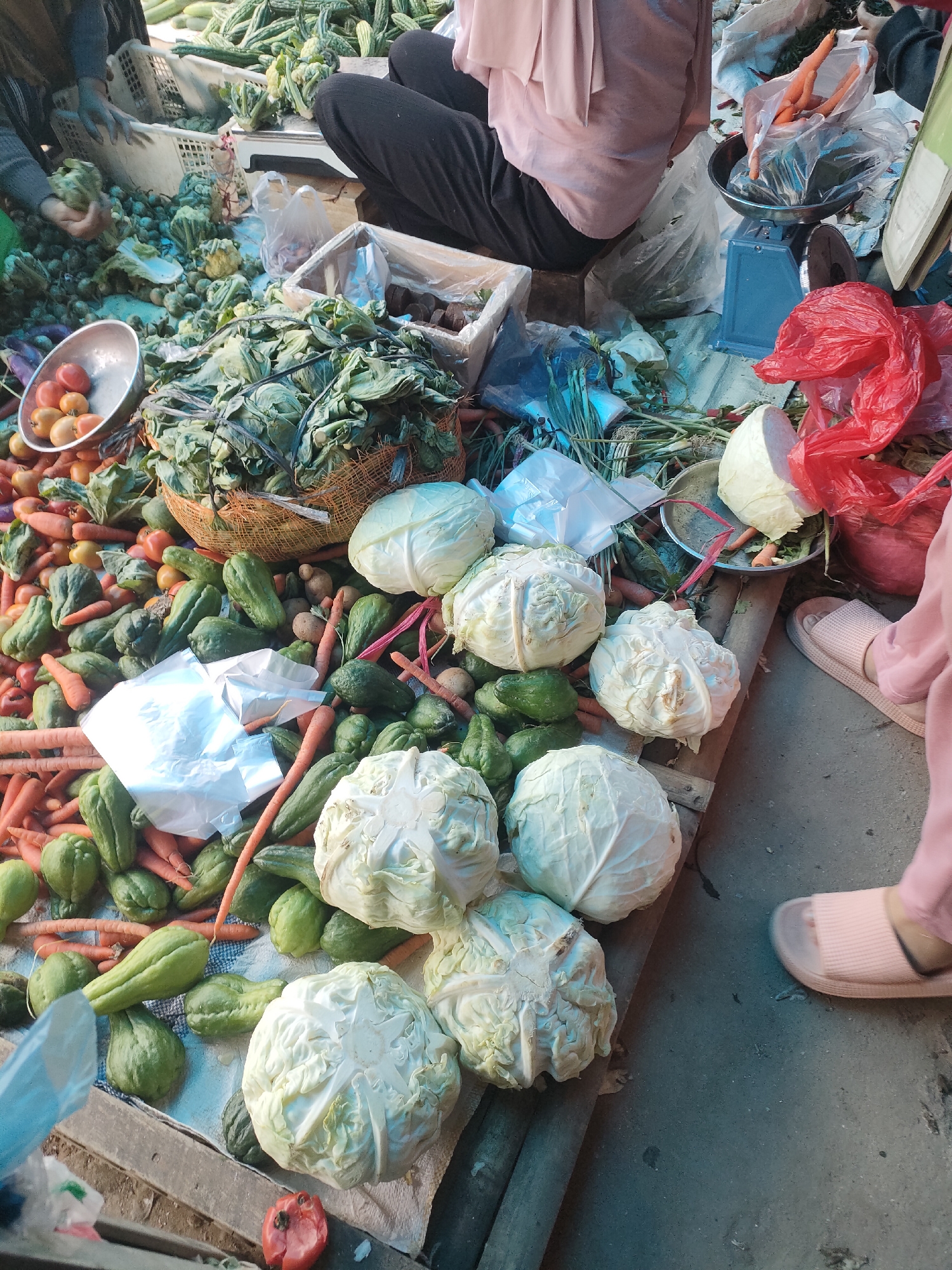 This image depicts an outdoor market stall, likely in Sukoyoso, Indonesia, overflowing with a variety of fresh produce. The scene is dominated by piles of vegetables, including numerous white cabbages, green chayote squash, and bright orange carrots. Several bunches of leafy greens are also visible, some contained within a woven basket.
In the background, a person wearing black pants and a light pink top is seated, interacting with the produce. Another individual, partially visible on the left, appears to be sorting vegetables. A blue mechanical scale is positioned on the right, suggesting items are being weighed for sale. Red plastic bags and white plastic bags are scattered throughout the stall, containing or holding vegetables. The ground surface appears to be concrete, and the overall impression is of a busy, active market environment.