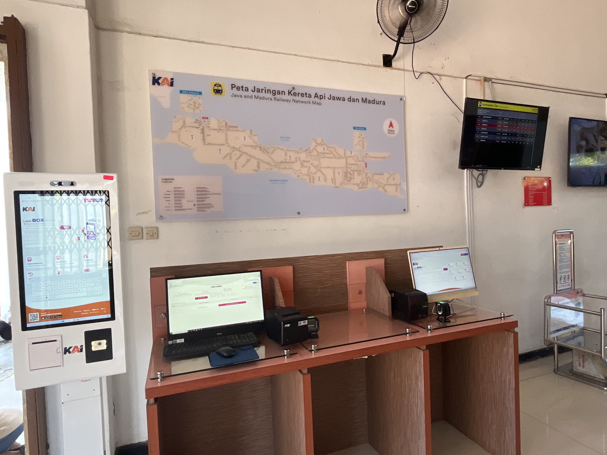 The image shows a ticket counter and information display area within what appears to be a train station or transportation hub in Desa Tambakreja, Indonesia. On the left, a large white touchscreen kiosk labeled "KAI BOX" displays information, likely for ticket purchases or travel information. Next to it, a wooden counter houses two desktop computers with keyboards and mice, and printers. The computers display screens with what looks like booking interfaces. On the wall above the counter, a large map titled "Peta Jaringan Kereta Api Jawa dan Madura" (Java and Madura Railway Network Map) is displayed. To the right of the map, two flat-screen televisions are mounted on the wall; one shows a grid of information, likely train schedules, and the other displays a scenic image. A ceiling fan is visible in the upper right corner. The environment is clean and organized, with tiled flooring and neutral-colored walls. No individuals are clearly visible in the foreground or actively engaged with the equipment, though a person's leg is partially visible in the lower left corner.