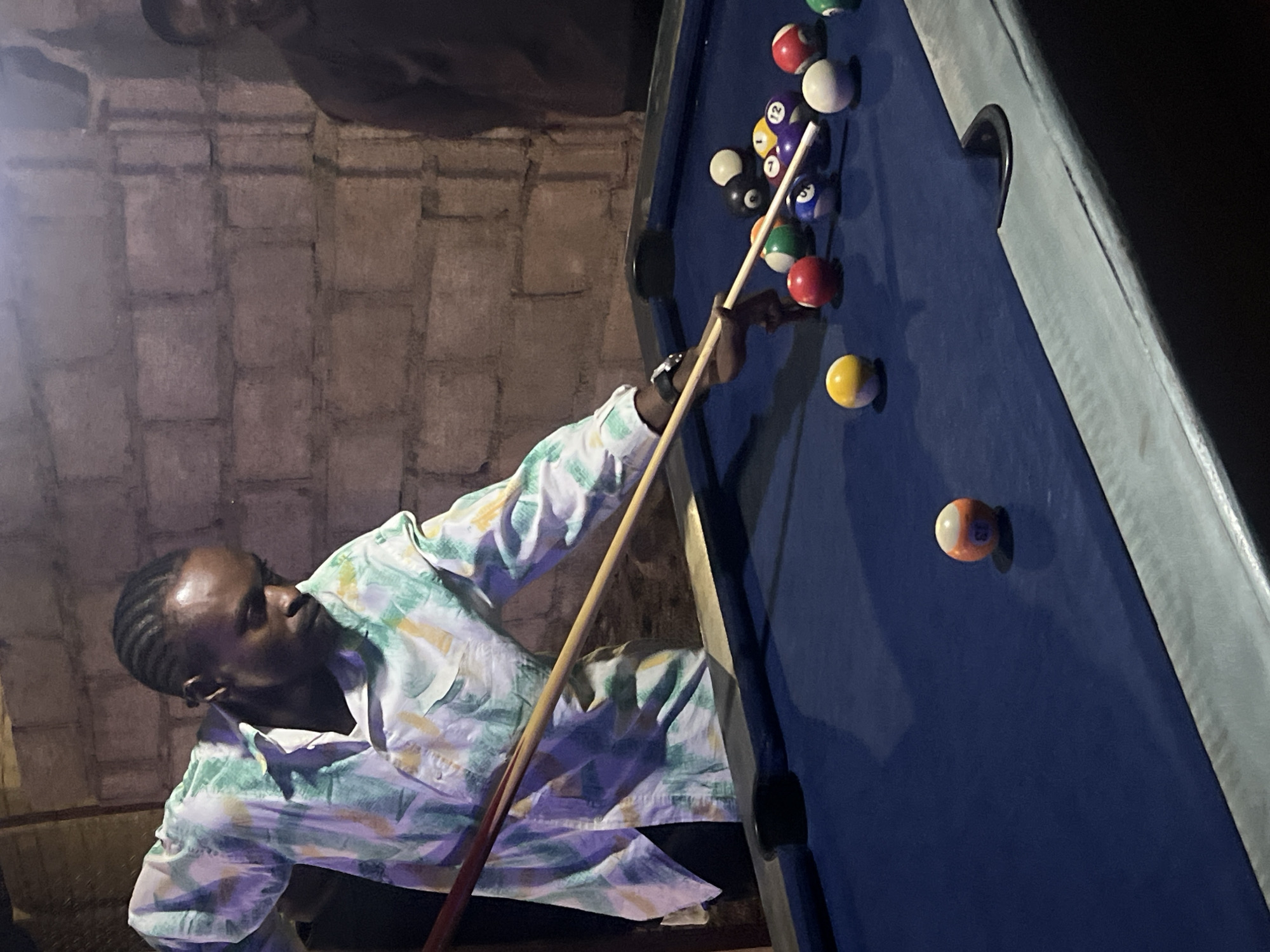 A man with braided hair stands at a blue pool table, cue in hand, preparing to strike a cluster of billiard balls. The backdrop is a weathered, gray brick wall. The scene is dimly lit.