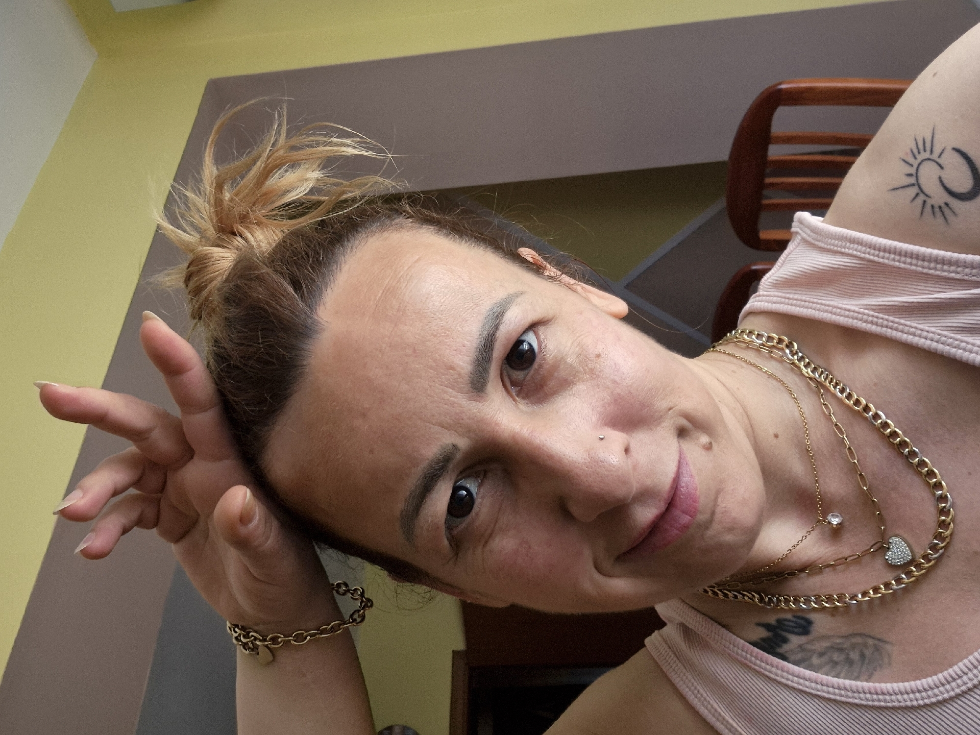 A woman with brown hair pulled into a messy bun is the central subject. She has fair skin, brown eyes, and a piercing on her nose. She is smiling slightly. She is wearing a pink tank top and several gold necklaces. She is also wearing a gold bracelet. There is a tattoo on her arm of a sun and moon. The background consists of a yellow and brown wall, a wooden chair, and a dark doorway. The location is Unknown, Unknown.