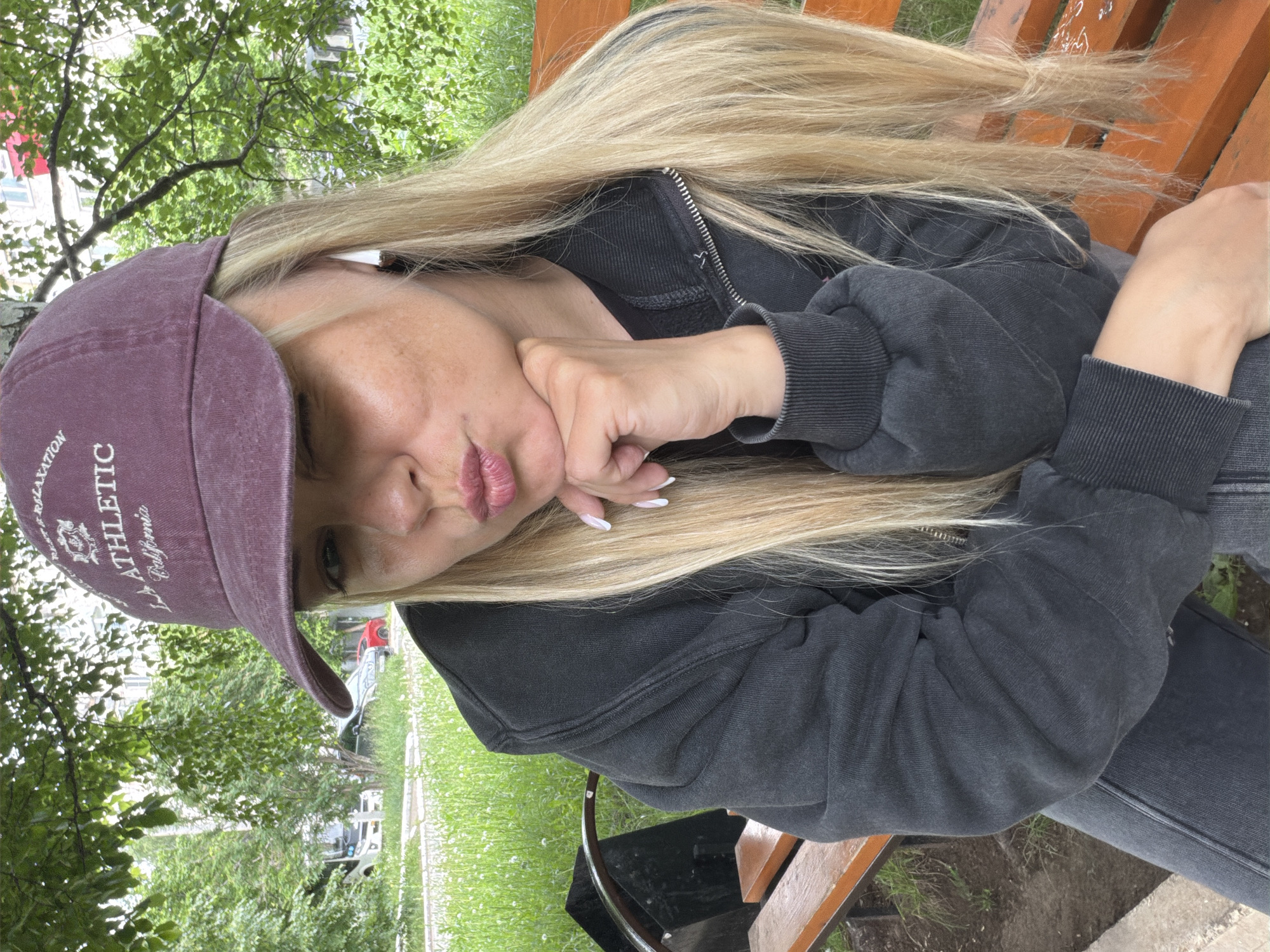 A young woman with long, straight blonde hair is captured outdoors, seated on a wooden bench in Votkinsk, Russia. She is wearing a dark grey zippered hooded sweatshirt and dark pants, paired with a maroon baseball cap. The cap features white embroidered text that reads "RELAXATION," "LA ATHLETIC," and "California." Her right hand, showing manicured nails with white tips, rests under her chin while she makes a playful "duck face" expression, puckering her lips.
The background is filled with lush green trees and grass, suggesting a park or a green area within a residential setting. Faint outlines of buildings are visible behind the foliage. The lighting indicates it is daytime, possibly under an overcast sky or in shade. A wooden bench she is sitting on is visible, along with a partial view of a dark metal object, possibly a chair or table base. The woman's face shows some visible skin texture. The image itself is rotated counter-clockwise.