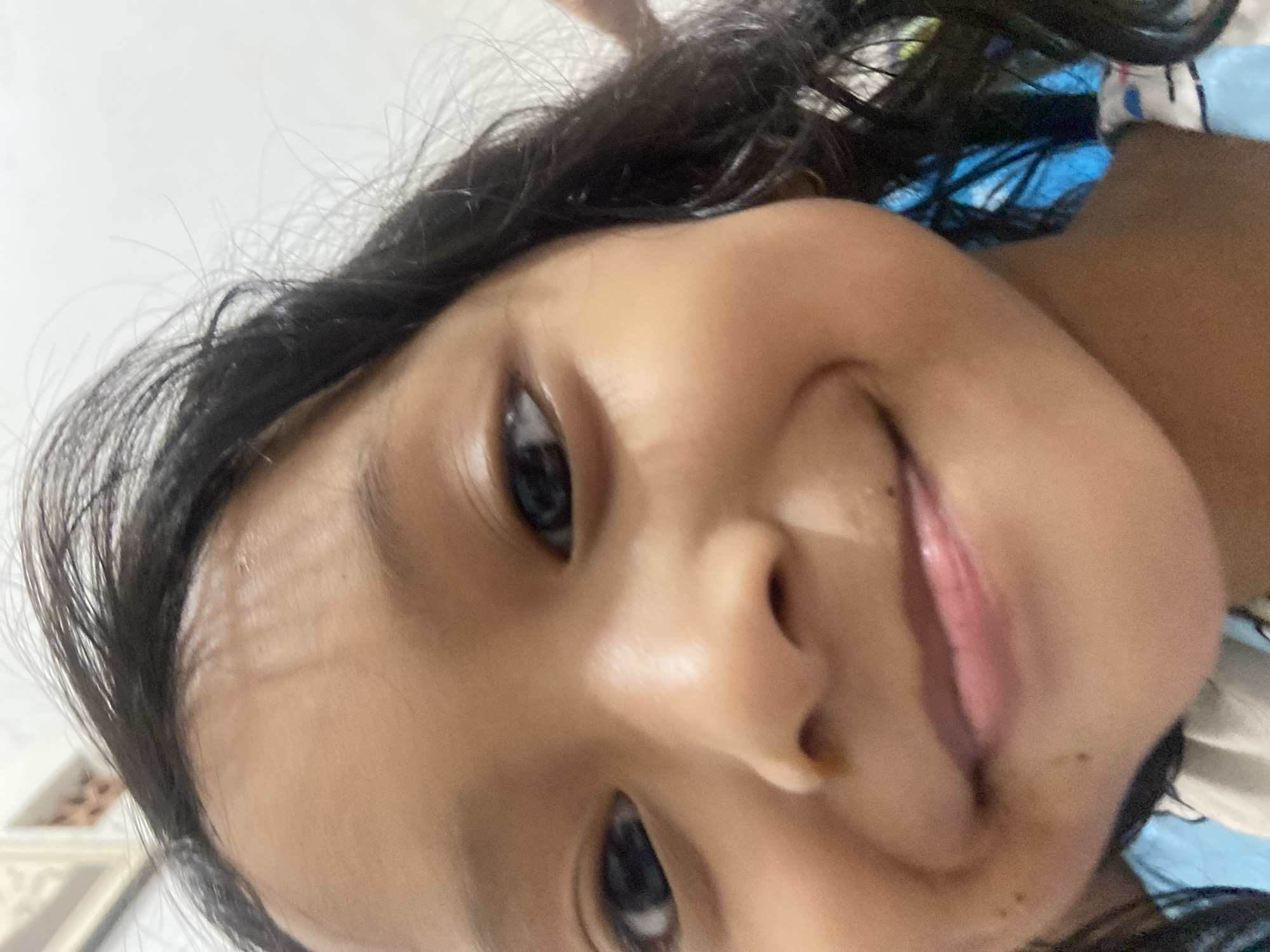 A close-up shot shows the face of a young girl, likely of Asian descent, with dark hair. The girl is looking directly at the camera and smiling, with her lips slightly parted to reveal a pink tongue. Her eyes are dark and wide, and her eyebrows are dark and well-defined. The background is out of focus and appears to be an indoor setting, possibly a room with a light-colored wall and a framed picture. The overall impression is one of a candid and happy moment captured.