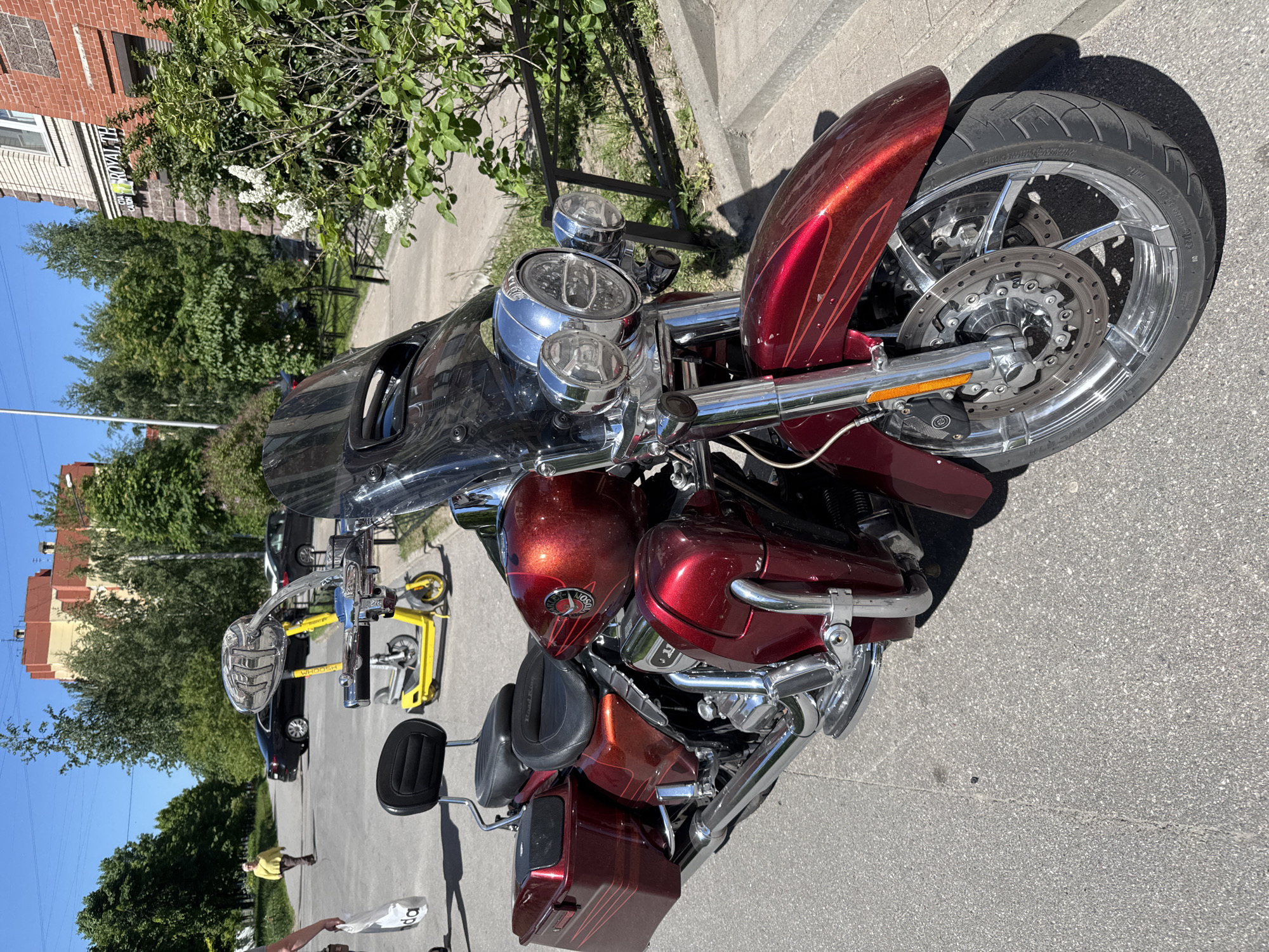 A shiny, dark red metallic flake motorcycle, identified as a Harley-Davidson Road King Special, is prominently parked on an asphalt street during a bright, sunny day. The motorcycle features extensive chrome accents on its handlebars, triple headlights, engine, exhaust pipes, and custom multi-spoke wheels with large disc brakes. Its paint job includes subtle pinstripes. A black leather seat with a backrest and matching saddlebags are visible. Text "WHOOEM" is readable on the motorcycle's side mirror.
In the background, to the left of the motorcycle, a black car and two yellow rental scooters are parked. Further back, several lush green trees and bushes, some with white flowers, line the street. A multi-story brick building with white windows stands behind the greenery, displaying a partial sign that reads "ROYAL TH". The sky is clear blue.
Two individuals are visible in the scene: one person in a yellow shirt walks in the mid-ground, holding a white shopping bag with the letters "da" printed on it. Another person's legs and a white shopping bag are visible in the foreground, close to the motorcycle. The strong sunlight casts distinct shadows across the asphalt and from the motorcycle, indicating mid-day. The setting appears to be an urban street in Saint Petersburg, Russia. The overall mood is calm and everyday.