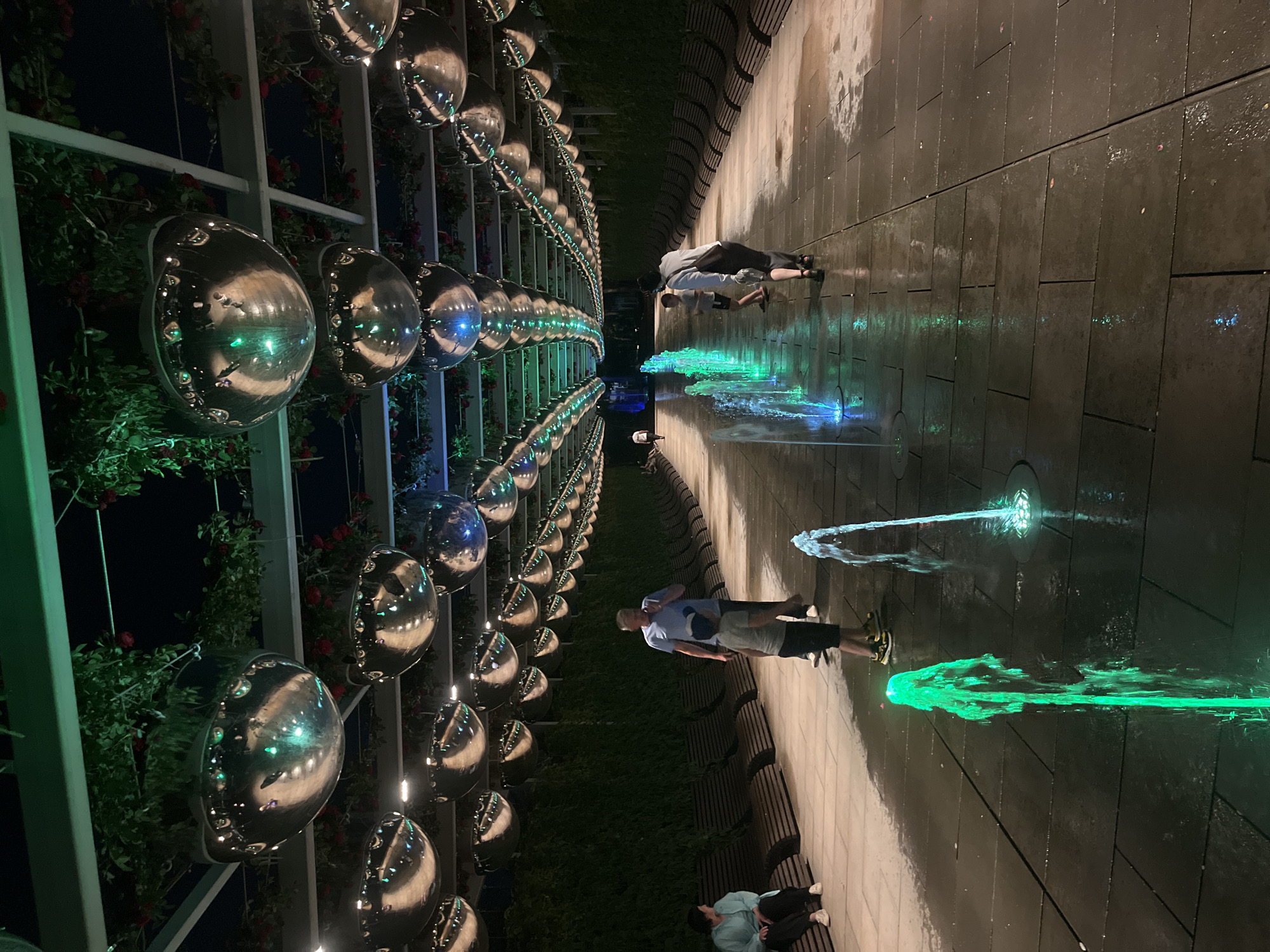The image depicts a night scene in the city of Krasnodar, Russia, likely at the "Chistye Luga" park, a popular recreation area known for its modern design and attractions.
In the foreground, a man and a child are walking near a series of illuminated fountains. The fountains are spraying water in jets, some of which are colored green and blue, reflecting on the wet paving stones. The man is wearing a blue t-shirt and dark shorts, while the child is in a grey t-shirt and dark shorts.
Further down the pathway, another man in a blue t-shirt and dark shorts is standing, possibly talking to a child who is out of frame. To the left, a woman in a light blue top and black pants is seated on a wooden bench. Behind the benches, a dense green hedge lines the walkway.
The walkway itself is paved with large rectangular stone tiles, which appear wet and reflective, indicating recent watering or rain. Overhead, a canopy structure is visible, from which numerous large, mirrored spheres are suspended. These spheres reflect the ambient light, creating a dazzling visual effect. Red flowers and green foliage are draped along the beams of the canopy, adding a natural element to the artificial lighting and structures. The overall atmosphere is one of a vibrant, illuminated public space at night.