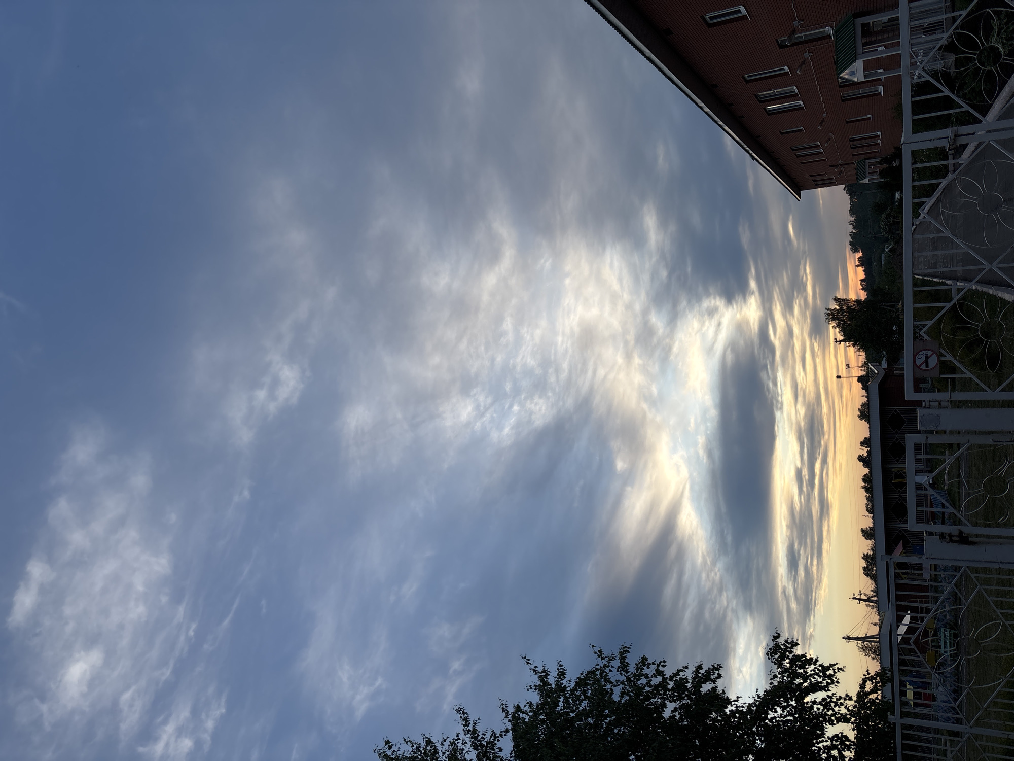 The image shows a cloudy sky at dusk. The lower portion of the image contains the top of a tree and a metal fence. Above the fence, there is a glimpse of a building, power lines, and other trees. The top of a red brick building is partially visible in the upper right corner of the frame. The location is in the city of Фоки, Russia.
