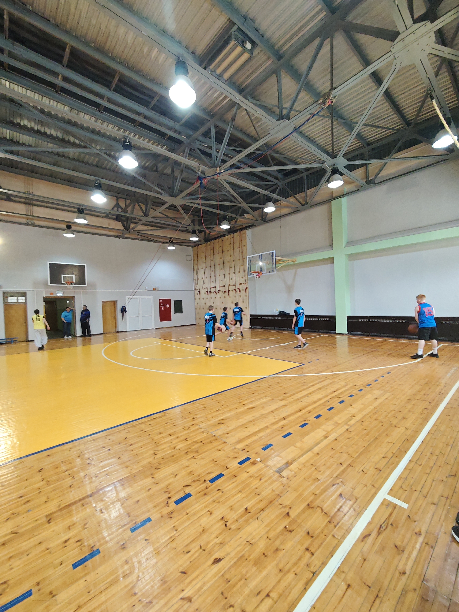 The image shows a basketball game in progress within an indoor gymnasium in Markovskii, Russia. Players wearing blue and black jerseys are actively engaged in the game. A player in a yellow jersey stands near the basketball hoop with spectators standing behind them. The court features a yellow free-throw lane, and the floor is light brown wood. The gymnasium has a high ceiling with exposed metal beams and numerous lighting fixtures.