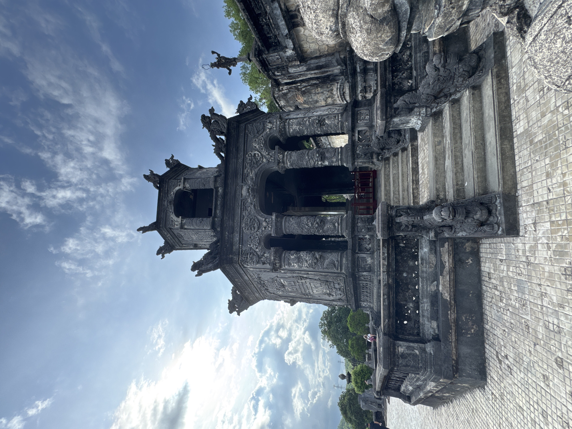 The image shows the exterior of a large, ornate stone structure, likely a tomb or temple, under a partly cloudy sky. The structure is dark gray and elaborately decorated with intricate carvings of dragons, mythical creatures, and geometric patterns. It features a multi-tiered roof with dragon figurines adorning the edges and a central octagonal tower. A wide set of stone stairs leads up to an open entrance flanked by columns. The ground in front of the structure is paved with small, diamond-shaped tiles. To the right, a large, weathered stone sphere is visible. In the background, trees and a distant power line can be seen. Two people are present in the scene: one person wearing a pink shirt and a backpack stands on the paved area to the left of the main structure, and another person is partially visible on the far left edge of the image. The overall atmosphere is solemn and historic. This structure is located in Thủy Xuân Ward, Vietnam, and is likely part of the Imperial Tombs of Huế.