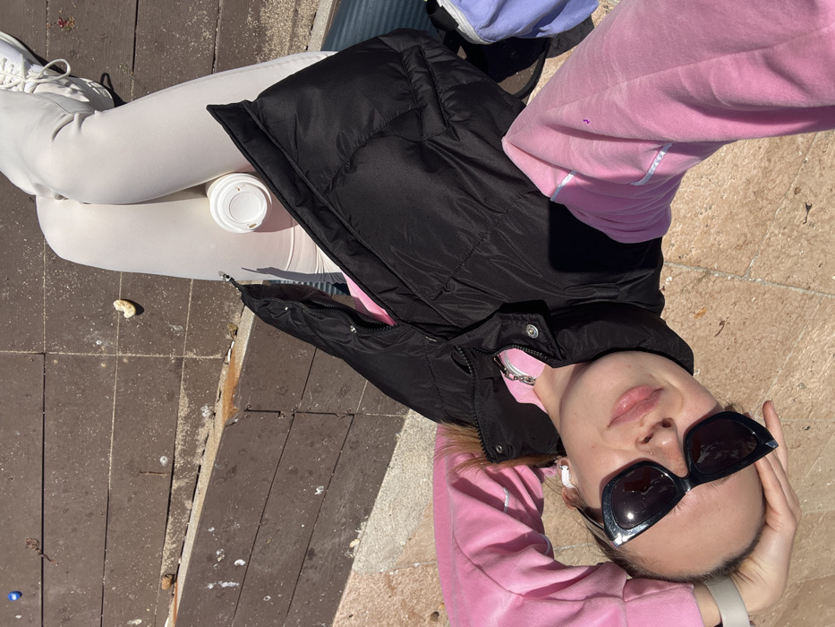 An overhead selfie captured in Budva, Montenegro, depicts a young female lying on her back, oriented diagonally with her head towards the upper right and feet towards the lower left. She wears a pink long-sleeved hooded top, a black puffy sleeveless vest, light-colored pants, and white sneakers on her left foot. Dark, cat-eye style sunglasses are worn, and white wireless earbuds are visible. Her left hand is positioned behind her head, with a light-colored wristband or watch visible. A white paper cup with a lid rests on her lap between her legs. A portion of a light blue bag is visible on the ground to her left. The ground consists of two adjacent sections: a light-colored, possibly tiled surface under her upper body, and a darker brown wooden boardwalk under her lower body. The wooden planks exhibit wear and are scattered with sand and small debris. Strong sunlight illuminates the scene, casting defined shadows.