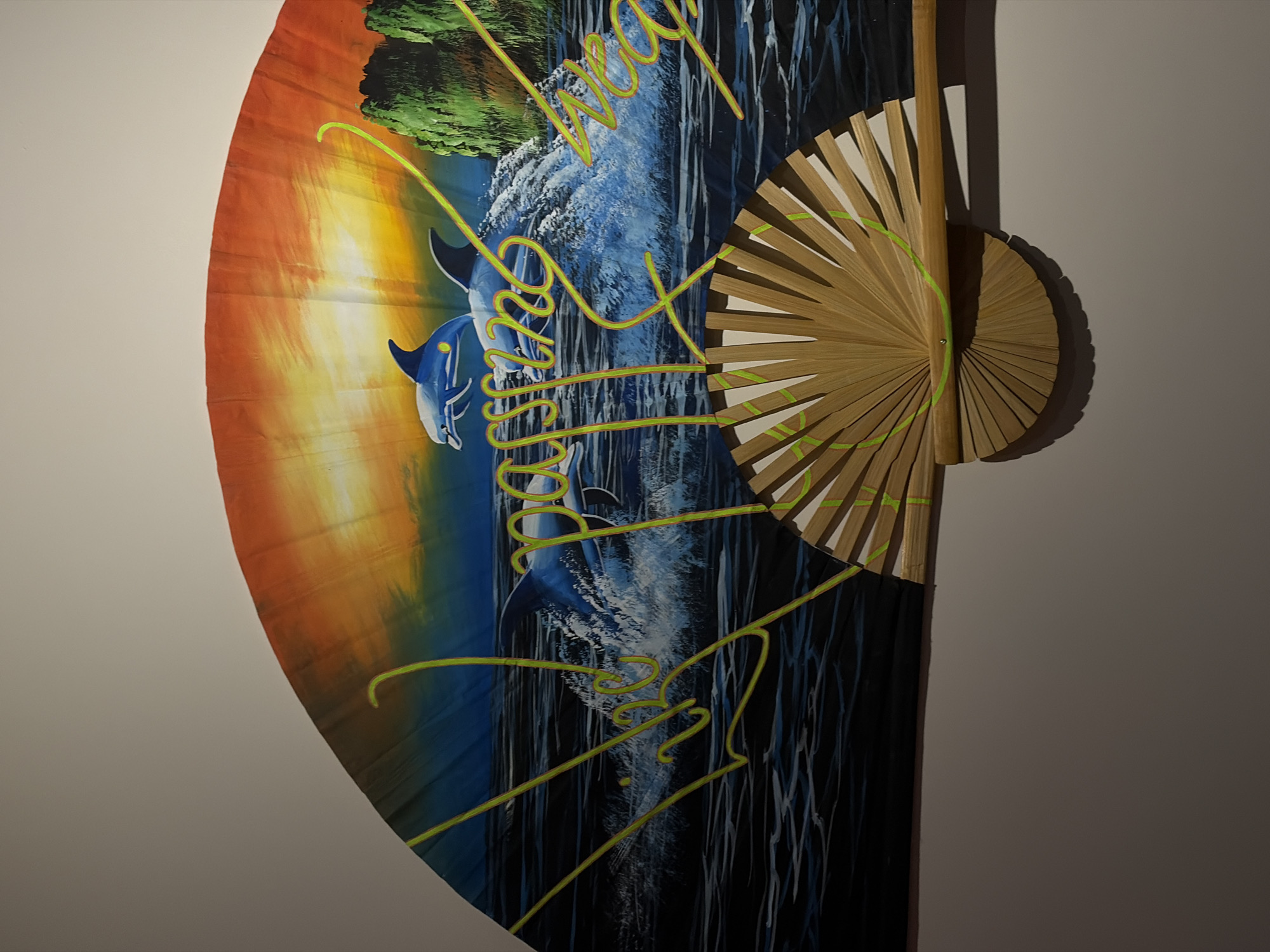 A decorative hand fan hangs on a plain wall. The fan is partially open, revealing a painted scene of dolphins jumping out of the water against a backdrop of sunset hues blending into a stormy ocean. The fan's ribs are made of light-colored wood, with some accented with lime-green markings. Calligraphic text in a similar lime-green is overlaid across the painted scene. The image captures the fan in its entirety, showcasing the detailed painting and the bamboo structure. The overall impression is of a decorative piece that may have been purchased in a tourist area. Given the imagery of tropical ocean wildlife, it could be associated with coastal regions near Bangkok, or more likely, available for tourists in urban markets. The wall is without any other decoration, drawing the full focus on the fan.