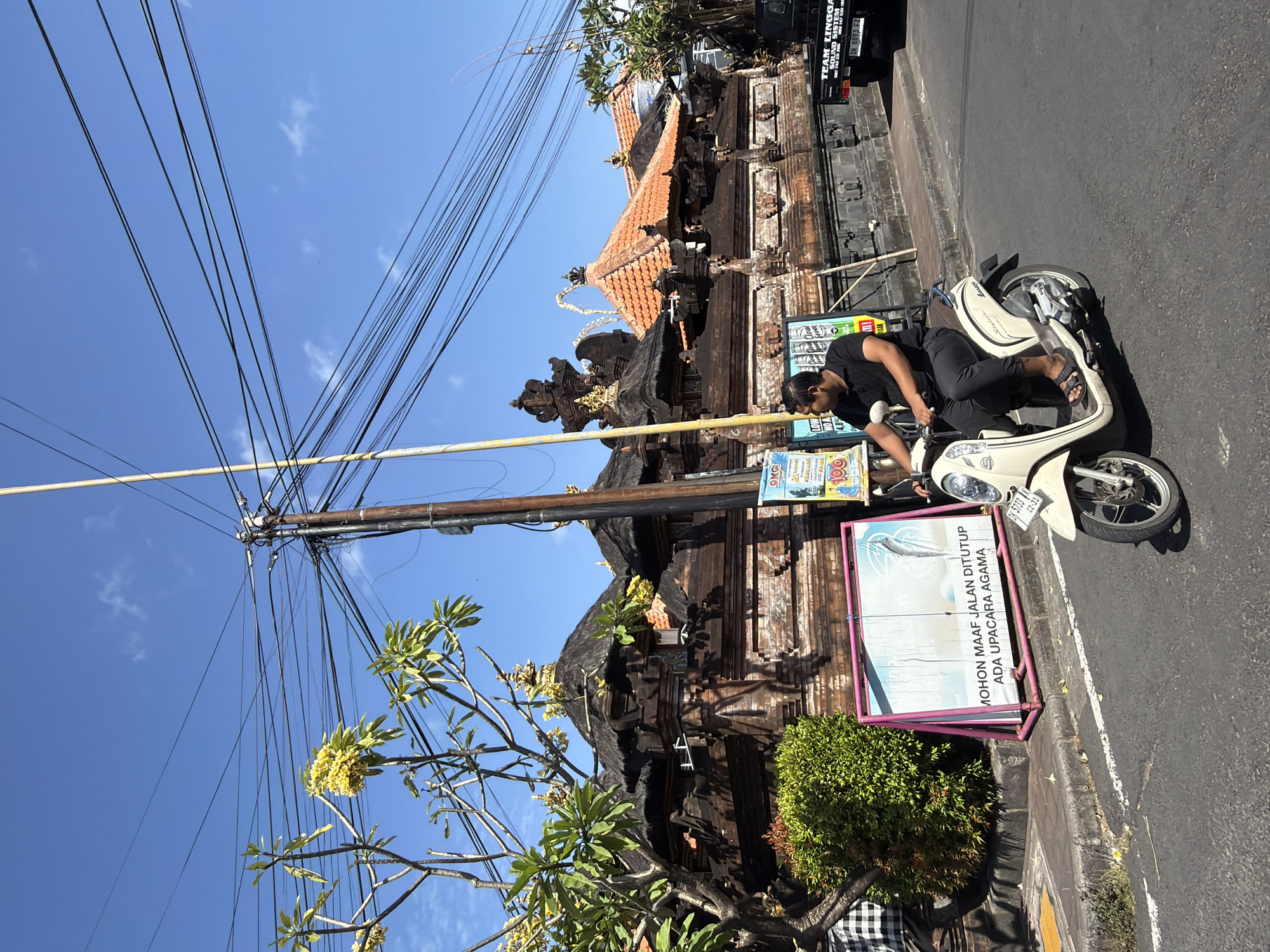 A man rides a light-colored scooter down a paved road in Kuta Selatan, Indonesia, on a clear, sunny day. He is wearing a black polo shirt and black pants, with black sandals. The scooter is positioned on the right side of the road. In the background, there is a traditional Balinese temple with dark, thatched roofs and intricate stone carvings. A large billboard, displaying text in Indonesian that reads "MOHON MAAF JALAN DITUTUP ADA UPACARA AGAMA" (Sorry, the road is closed due to a religious ceremony), is propped up on the sidewalk to the left of the temple entrance. Above the temple, numerous power lines crisscross the blue sky, supported by wooden utility poles. A tree with yellow flowers is visible to the left of the temple. A black truck with the words "TEAM LINGGA SOUND SISTEM" on the back is parked further down the road to the right.