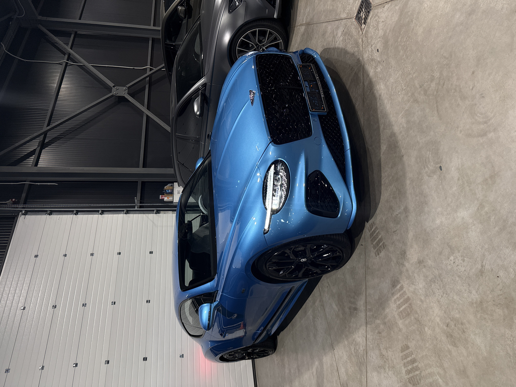 The image shows a blue Bentley Continental GT parked indoors. The car is the primary subject, positioned at an angle to showcase its front and side. It has a distinctive black grille and black wheels, and the Bentley logo is visible on the hood. A gray car is partially visible behind the Bentley, also parked indoors. The setting appears to be a garage or showroom, with a large white corrugated door in the background and a concrete floor. The lighting suggests it is either daytime or well-lit indoor conditions. There are some tire marks on the floor, indicating the car has been moved. No people are visible in the image. The license plate on the Bentley is not legible enough to read. The location is specified as Plovdiv, Bulgaria.
