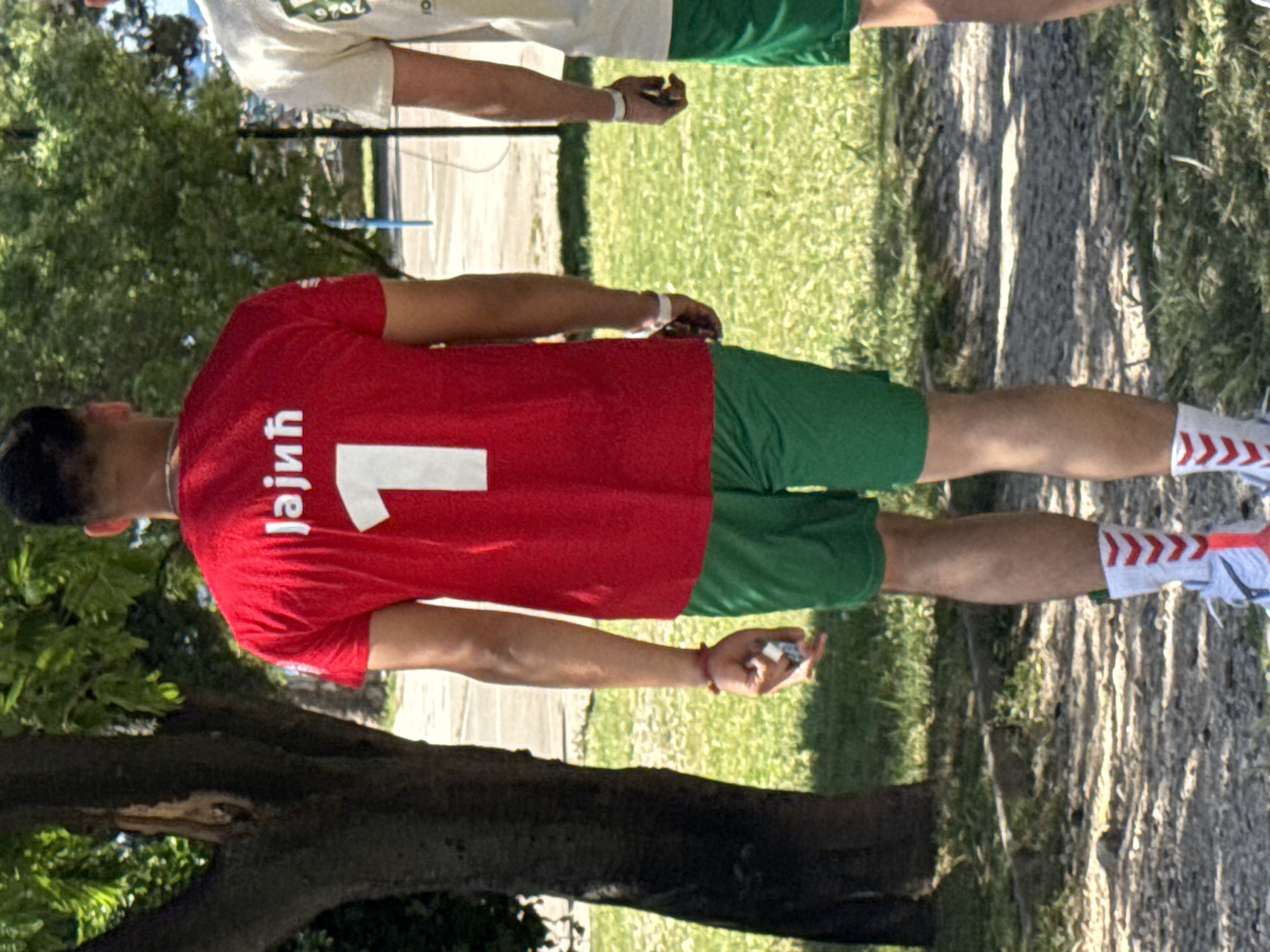 A medium shot captures two individuals outdoors. The first person wears a red t-shirt with the number 1 on the back and the name "Jajnih" in Cyrillic letters above it. They also wear green shorts and white socks with red chevrons near the ankles. This person is standing on grass near a tree trunk. The second person wears a light-colored shirt and green shorts. They are also standing on grass, with part of a pathway visible in the background. The location appears to be outdoors, likely a park or field, with trees and grass in the background. While the information is not explicitly available in the provided image, the city of Budva, Montenegro, is mentioned in the prompt.