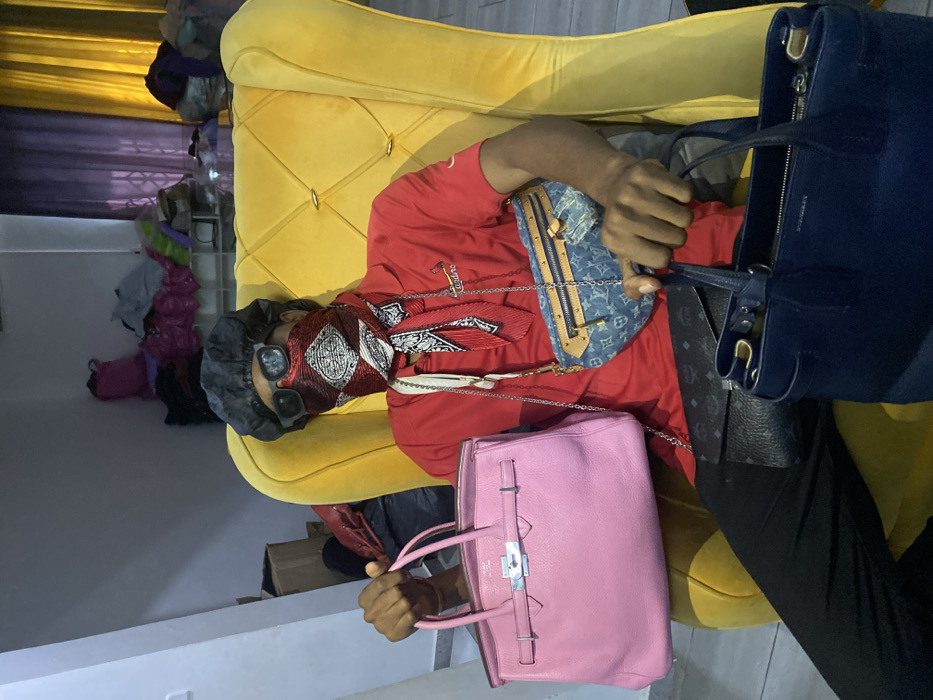 A single individual is seated in a prominent, tufted yellow armchair within an interior space, described as Tema, Ghana. The person wears a red collared shirt featuring an emblem, black trousers, and a black durag. Their face is partially concealed by dark sunglasses and a red and white patterned bandana covering the nose and mouth. A matching red and white patterned scarf is tied around the neck, and multiple silver-toned chain straps are visible, some connected to bags. The individual is actively holding a pink, pebbled-leather handbag, consistent with a Hermes Birkin style, in their left hand. A blue denim Louis Vuitton fanny pack with a chain strap is worn across the torso, and a black patterned clutch rests on the lap. A dark blue structured handbag, possibly Burberry, is held in the right hand. The background features light grey walls to the left and a darker, possibly purple, wall to the right. A white shelving unit displaying various piles of clothing or textiles is visible behind the armchair. Cardboard boxes are present on the left, and purple and yellow curtains are visible on the right side of the frame. The floor is tiled in grey.