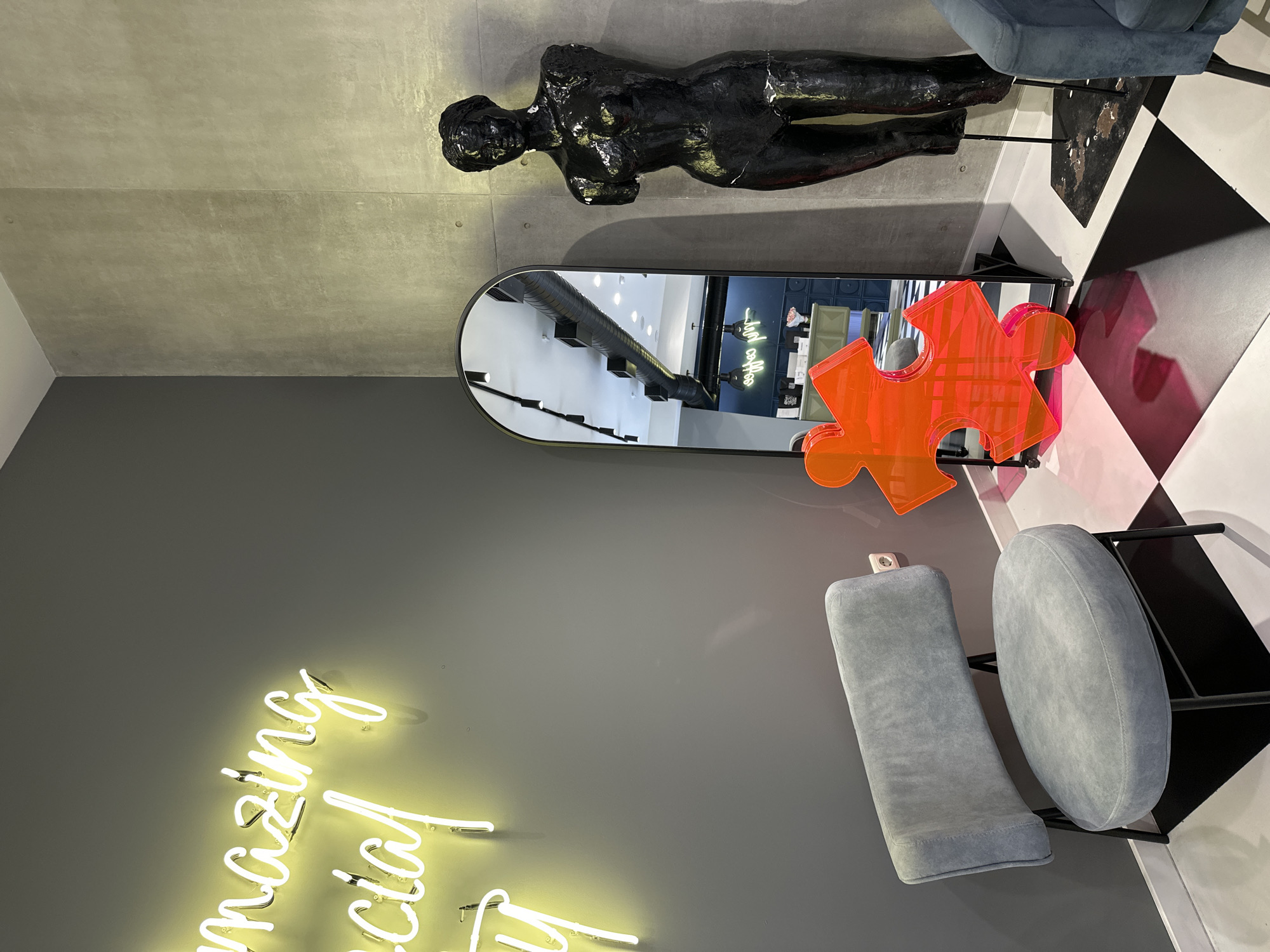 The image captures an interior scene with a peculiar artistic arrangement. On a gray wall, a neon sign spells out "amazing special day". Above this, a black full-bodied sculpture lies horizontally, seemingly supported by the wall. A sleek, elongated mirror reflects the interior of what appears to be a retail space, with visible signage and shelving. An oversized, bright orange puzzle piece sculpture is positioned near the mirror. Two plush, grey chairs sit near a black and white checkered floor. The scene is well-lit, with an emphasis on the contrasts between the textures and colors. The specific location in Yerevan, Armenia, cannot be determined from the visual information available.