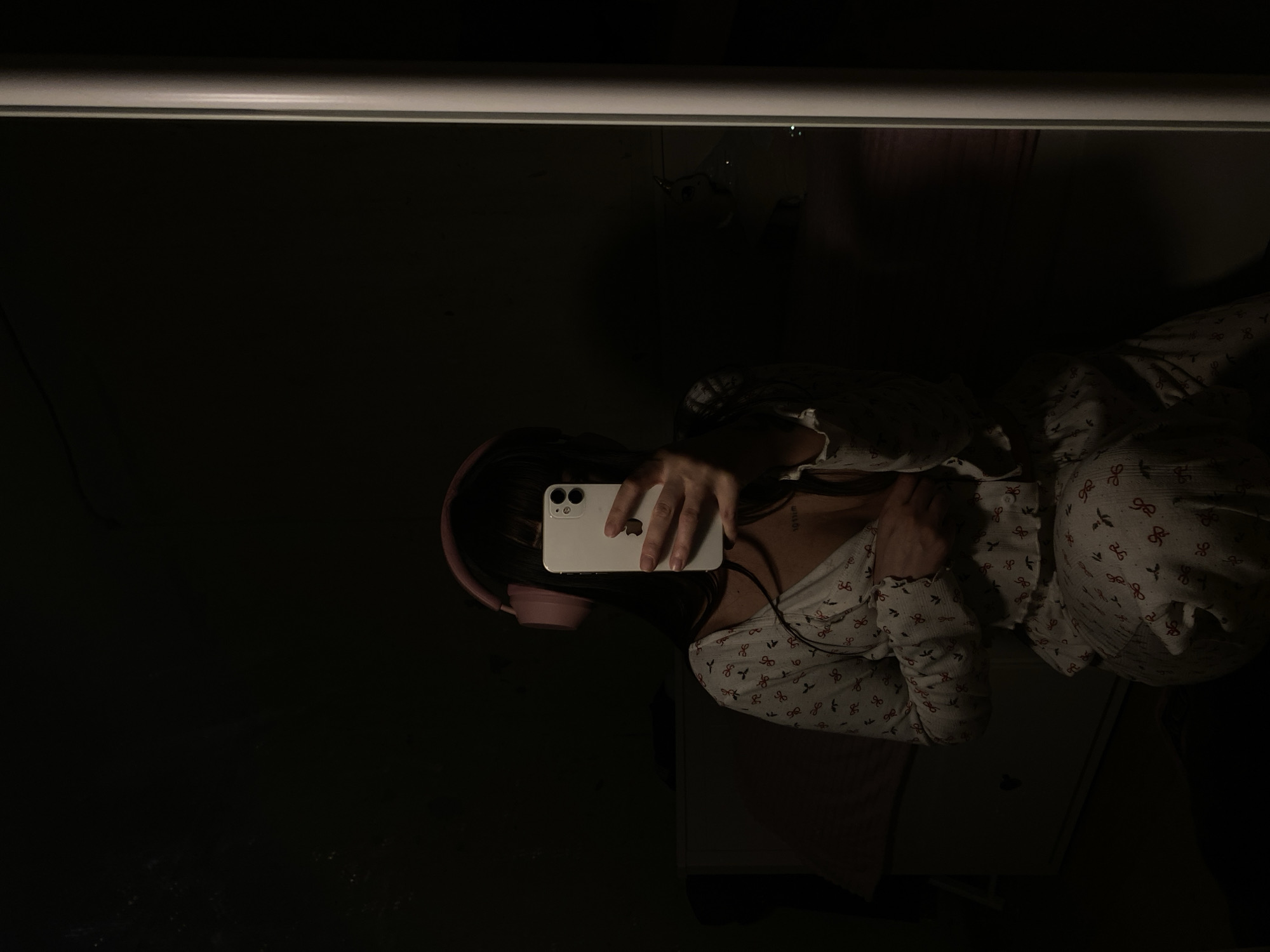 A person is visible in a dark environment, seemingly taking a photograph in a mirror. The person is wearing light-colored pajamas with a pattern of red bows and has pink headphones around their neck. They are holding a white smartphone in their hand, likely using it to take the picture. A small portion of a white piece of furniture is visible in the lower right. A light source illuminates the top of the frame. The lighting is poor, making details hard to discern. The exact location within the city of Votkinsk, Russia, is undeterminable from the image. No specific actions or interactions are evident beyond the person taking a photo.
