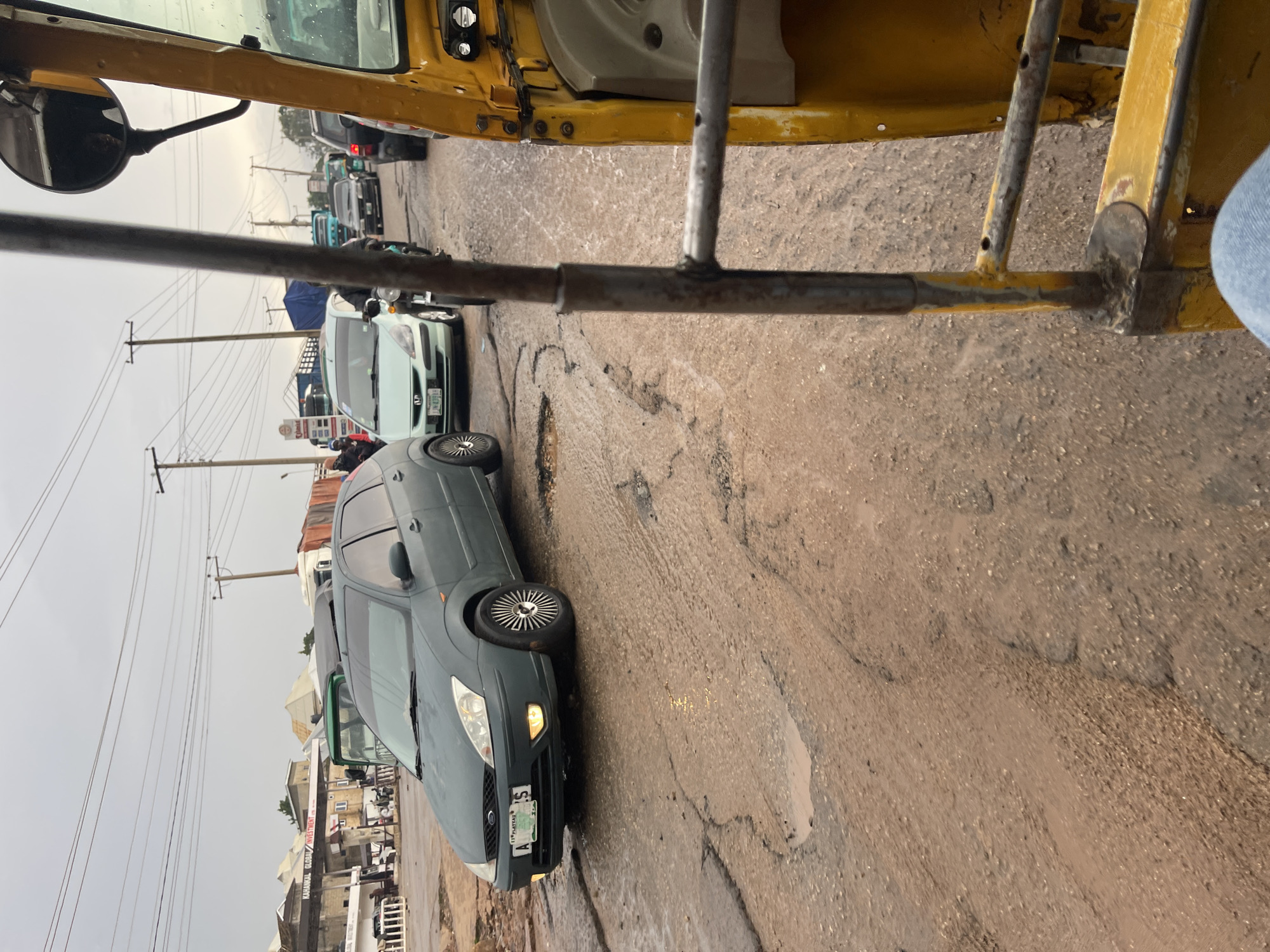 A dark green Ford Focus sedan is parked on a very broken and muddy road in Jos, Nigeria. The car is facing the left side of the image. The road is filled with potholes and puddles. In the background, there are several other vehicles, including a white minivan and a yellow auto-rickshaw which the photo is taken from. There are also buildings and utility poles visible in the distance. The sky is overcast, suggesting it may have rained recently. The scene appears to be a traffic jam or a busy intersection.