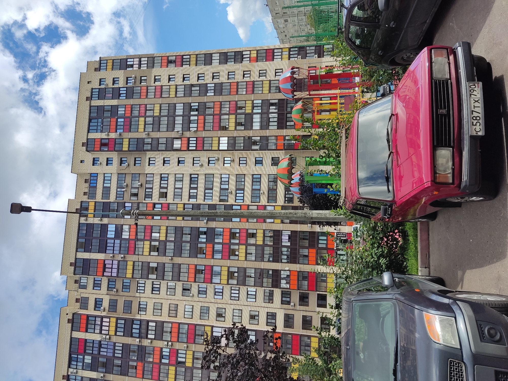 The image captures a bright, partly cloudy day in Noginsk, Russia, with a tall, modern apartment building dominating the background. The building features a light-colored brick facade with a rhythmic pattern of dark-framed windows and colorful balconies in shades of red, orange, yellow, and purple. A prominent streetlight stands to the right of the building. In the foreground, two cars are parked on an asphalt surface. The car on the left is a dark gray SUV, while the car on the right is a red sedan. A children's playground with colorful umbrella-shaped structures is partially visible between the cars and the building. Lush green bushes and trees are scattered throughout the scene, adding a touch of nature to the urban environment. The sky is a mix of blue and white clouds.