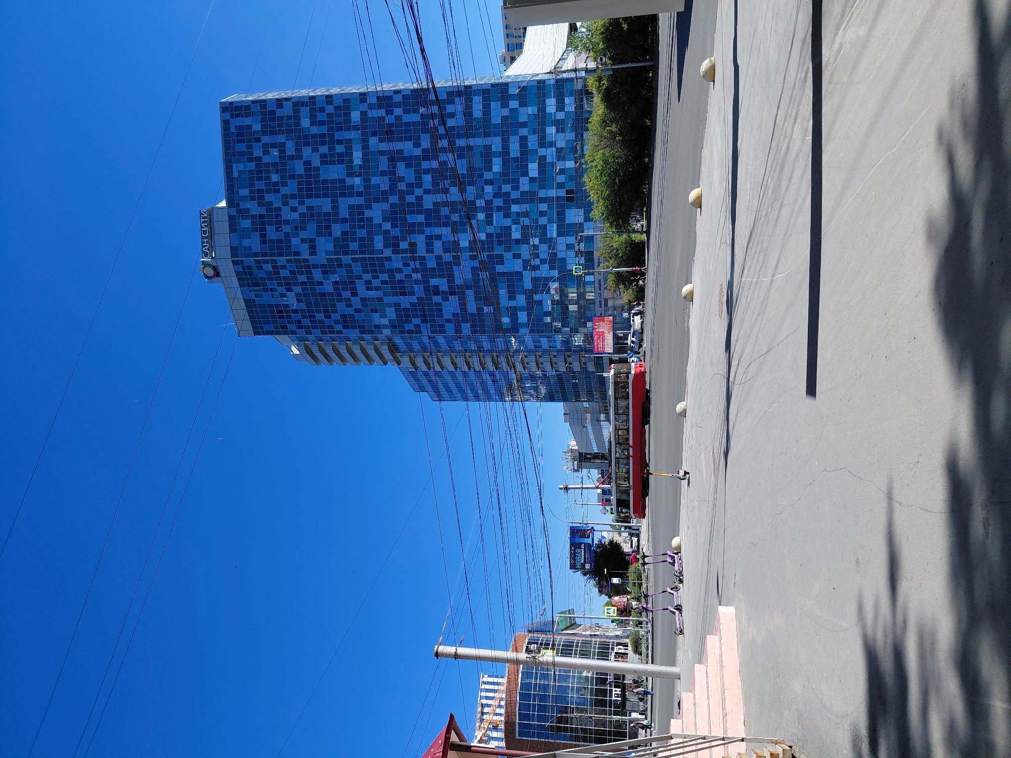 The media file depicts an urban street scene on a sunny day. The most prominent subject is a tall, modern building with a blue, tiled facade. To the right of the building is a paved street with a red tram or bus, and several parked electric scooters. There are also trees and other buildings visible in the background, suggesting a city environment. The sky is a clear, bright blue, and shadows indicate that the sun is high, likely mid-day.
In the foreground, there is a gray concrete sidewalk with a set of pink stairs leading up. The street is mostly empty of people, with a few figures visible near the tram/bus and in the distance. One notable detail is a logo or text on the blue building that reads "САН СИТИ" in Cyrillic script. Further down the street, another billboard is visible with text, though it's too small to be legible. The overall impression is a bright, clear day in a bustling urban area. There are no overt signs of specific events or strong emotions, but the scene conveys a sense of everyday urban life.