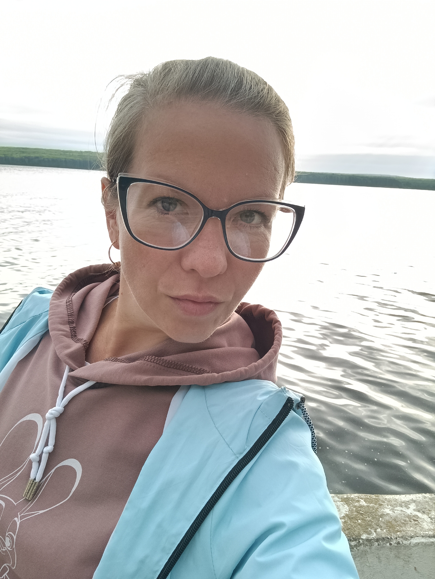 A woman with blonde hair pulled back and wearing glasses is taking a selfie outdoors. She is wearing a light blue jacket over a brown hooded sweatshirt with white drawstrings. Behind her, a body of water reflects the overcast sky, and a treeline is visible in the distance. The location is the Kama River embankment in Chaikovsky, Russia.
