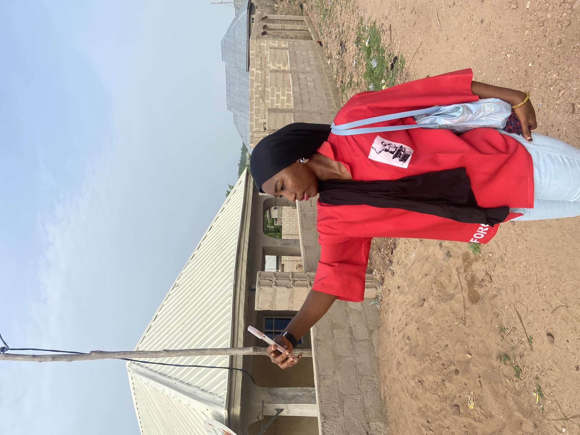 A young woman stands outdoors in Lokoja, Nigeria, holding a pen or similar object in her right hand, as if about to write or make a mark on something out of frame. She is wearing a red short-sleeved shirt with a black and white graphic on the front and the word "FOR" partially visible on the sleeve. The woman also has on light-colored jeans, a black head covering, and what appears to be a blue bag slung over her shoulder. She is looking down at the object in her hand.
Behind her are buildings, one with an incomplete block-like construction and the other with a corrugated metal roof. A utility pole with wires extends into the sky on the left. The ground is dry and sandy, characteristic of the environment.