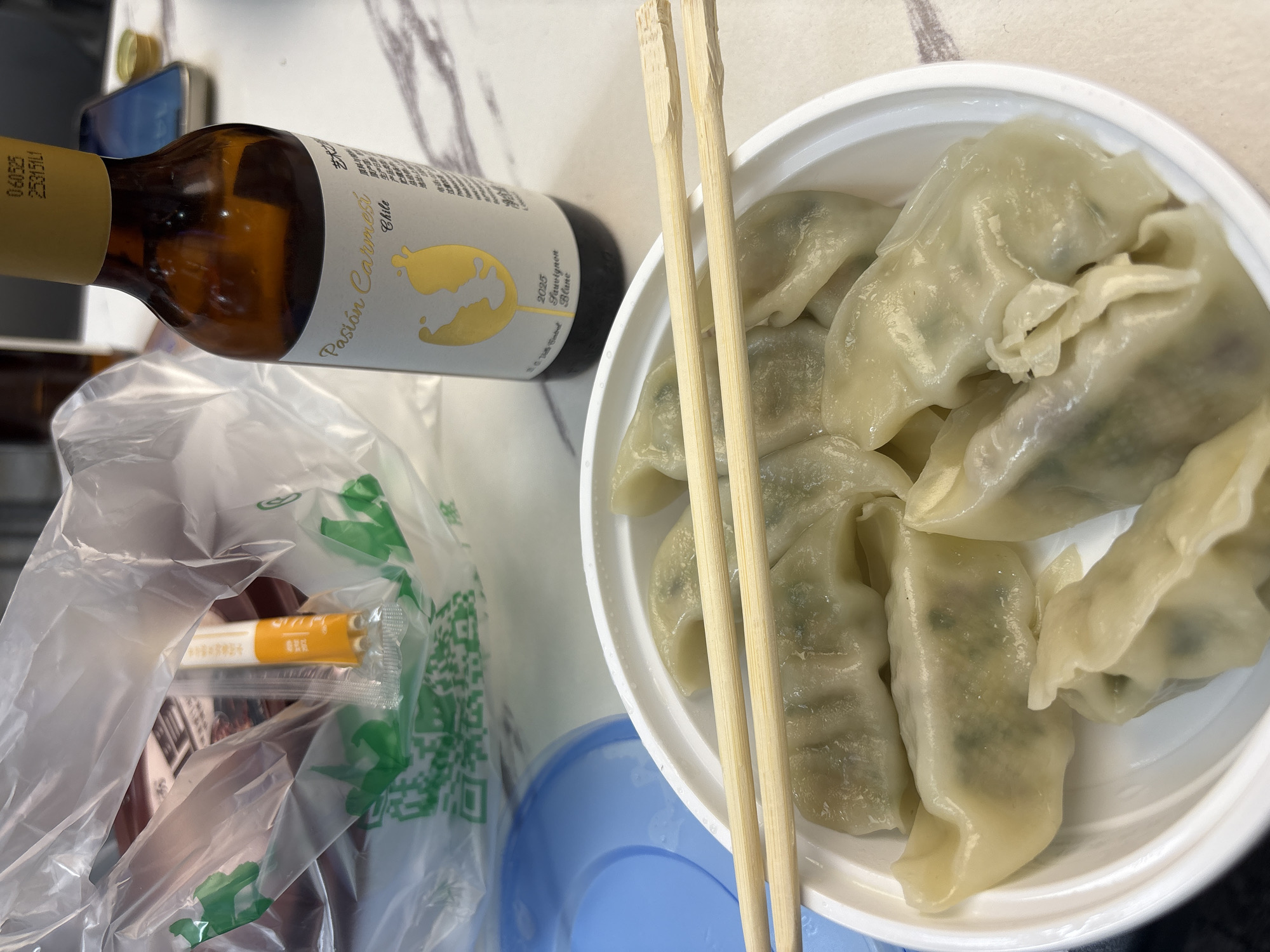 In a Haizhu District, China location, a close-up, eye-level shot presents a meal of dumplings in a white plastic bowl, accompanied by wooden chopsticks. The dumplings appear to be steamed, with visible green filling. To the right of the bowl, a small, amber glass bottle of wine with a yellow label is positioned. The label displays text in both English and Chinese, including "Pasion Carmeli Chile" and "2022 Sauvignon Blanc". Behind the bowl and bottle, a clear plastic bag is visible, containing other food items and a QR code printed on green packaging. A light blue plastic lid is partially visible to the left of the bowl. The surface appears to be a light-colored, patterned tabletop. The lighting is bright, casting soft shadows.