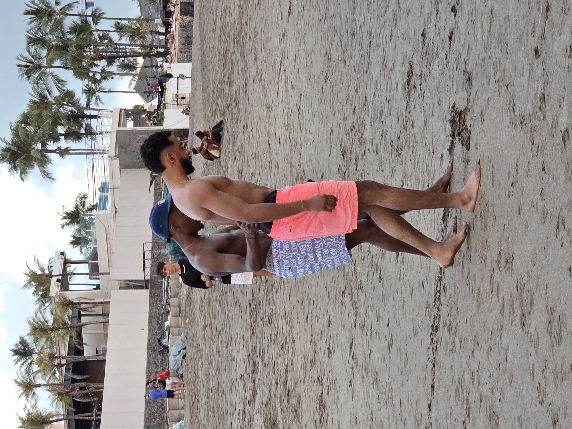 The image depicts a beach scene in North Kuta, Indonesia. In the foreground, two shirtless men are walking on the sand. The man on the left wears blue patterned shorts, while the man on the right wears bright pink shorts. Behind them, other individuals are visible on the beach, some sitting or standing. In the background, palm trees line the shore, and a white structure with multiple levels and balconies suggests a resort or restaurant. The sky is overcast. The sand is a light brown, with some debris and footprints scattered across it.