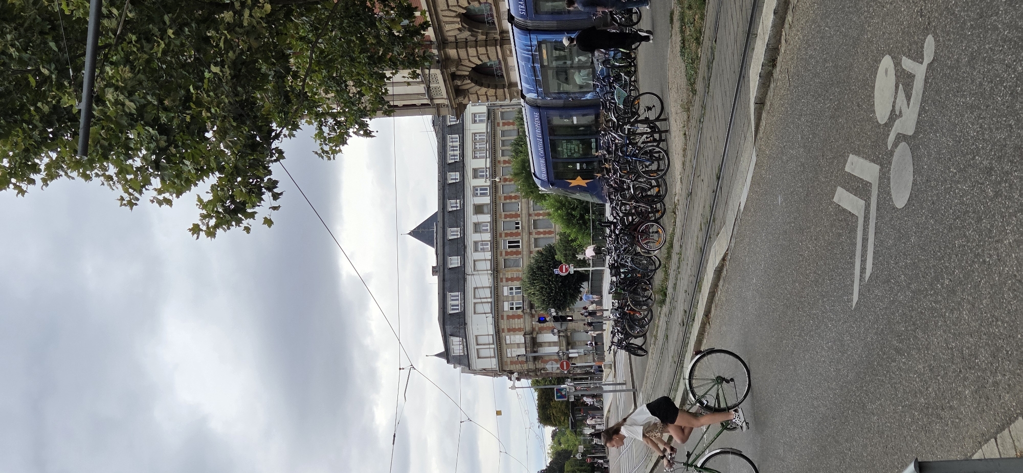This image captures a scene in Strasbourg, France, near a tram line and a building with a multi-story facade. The sky is overcast. In the foreground, a young woman with blonde hair is riding a bicycle on a street marked with a bicycle lane symbol. She is wearing a white t-shirt and dark shorts. Behind her, a large rack of parked bicycles is visible. Further back, people are walking on a bridge or elevated walkway. A blue tram with the text "STRASBOURG, CAPITALE EUROPÉENNE" and a yellow star is stopped on the tram tracks. Trees and overhead wires are present in the background.