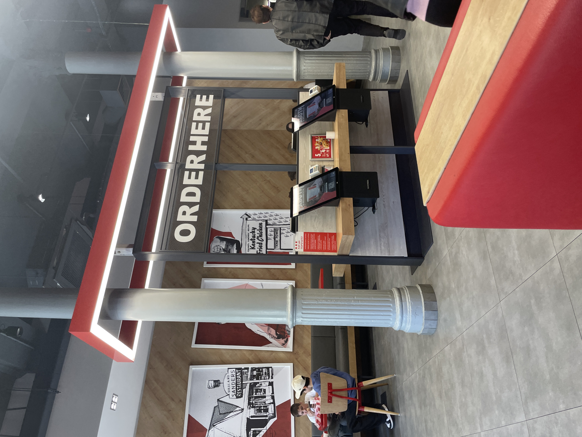 The image captures the interior of a KFC restaurant located in Plovdiv, Bulgaria. The central focus is a "ORDER HERE" sign structure. The restaurant features self-ordering kiosks with touch screen monitors and corresponding hardware. The decor includes framed prints related to KFC's history and Colonel Sanders. There are patrons in the dining area, including a man standing near the ordering station and a group of people seated at a table. The overall setting is a modern fast-food establishment.